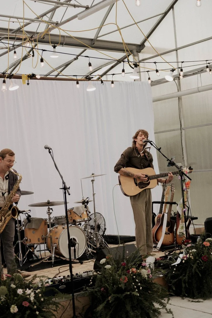 A band of musicians performing on stage inside a tent with string lights overhead. One musician is playing a guitar and singing into a microphone, another is playing drums, and there are additional guitars and floral decorations at the front of the s