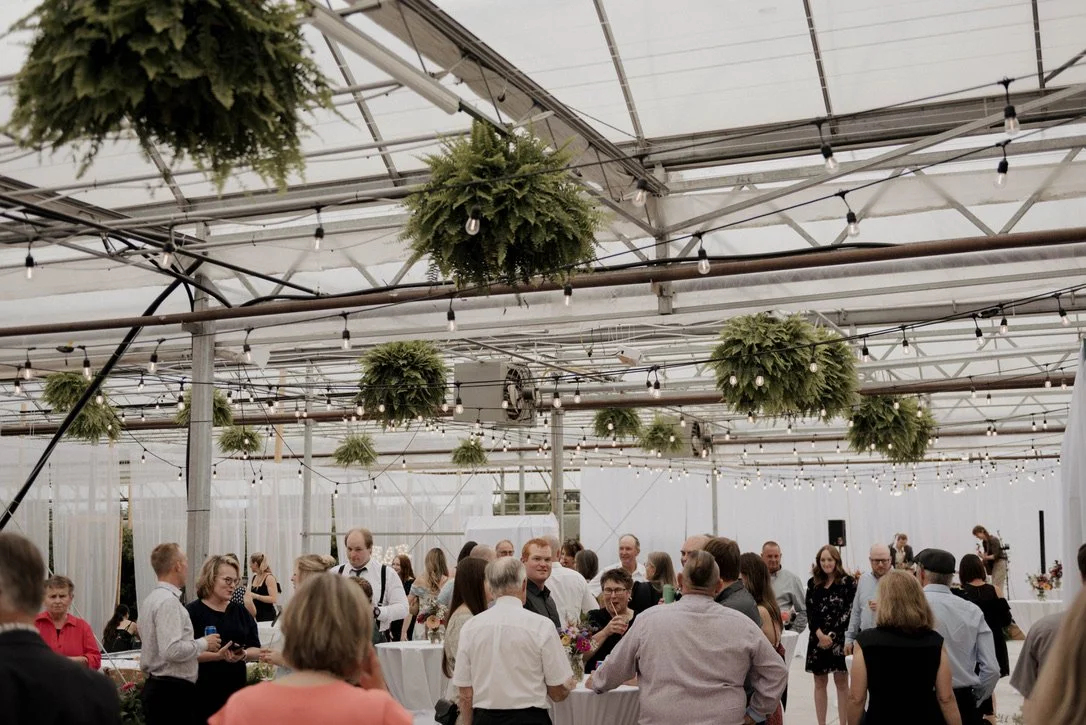 People gathered at a social event or party inside a greenhouse or green building with hanging plants and string lights.