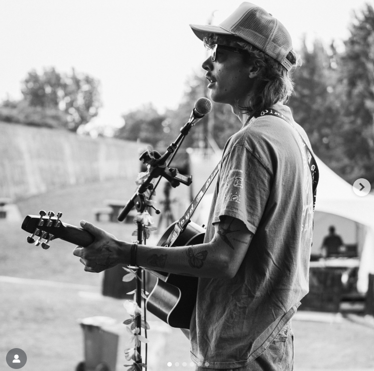 A young man wearing sunglasses, a cap, and a t-shirt playing an acoustic guitar and singing into a microphone at an outdoor event.