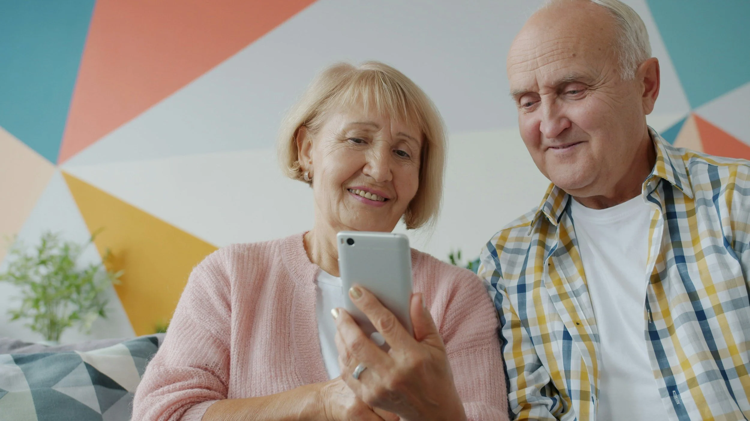 An older woman and man sitting together, looking at a smartphone with smiles. The woman is holding the phone. They are in a room with a colorful geometric wall and some potted plants.