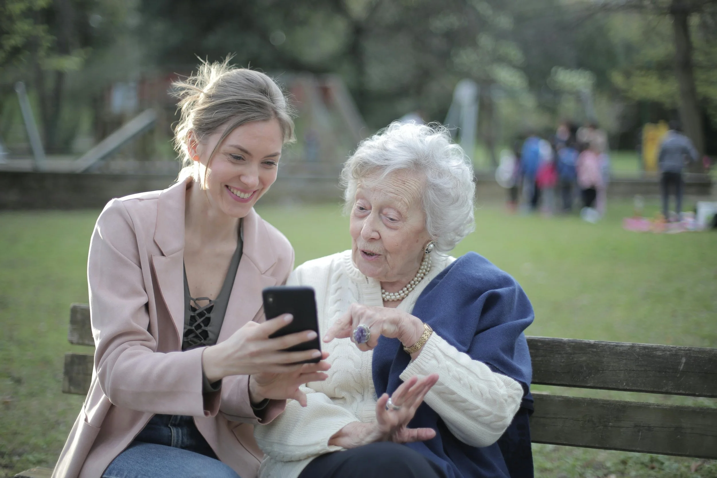A young woman and an elderly woman sitting on a park bench looking at a smartphone, smiling and engaging with each other.