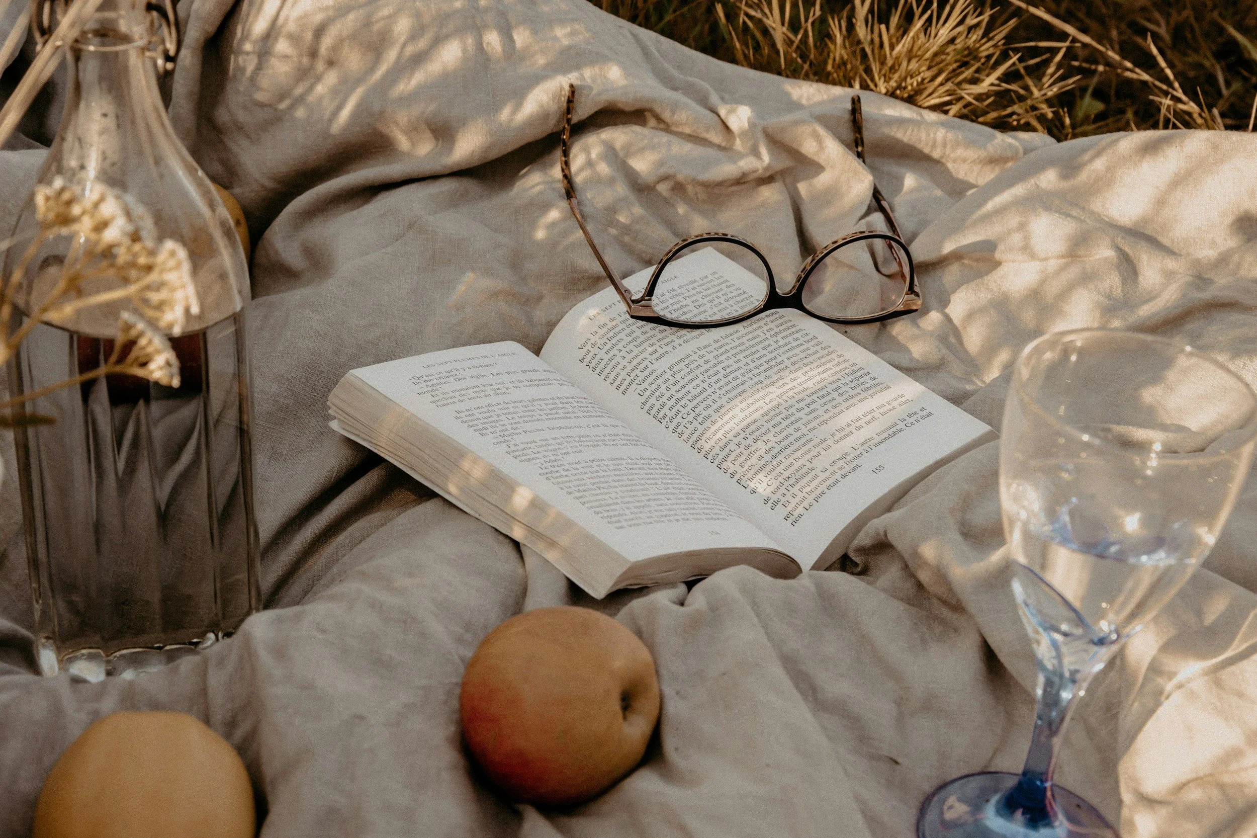 An open book with reading glasses on top, a glass of water, a vase with dried flowers, and two peaches resting on a soft light-colored fabric surface.