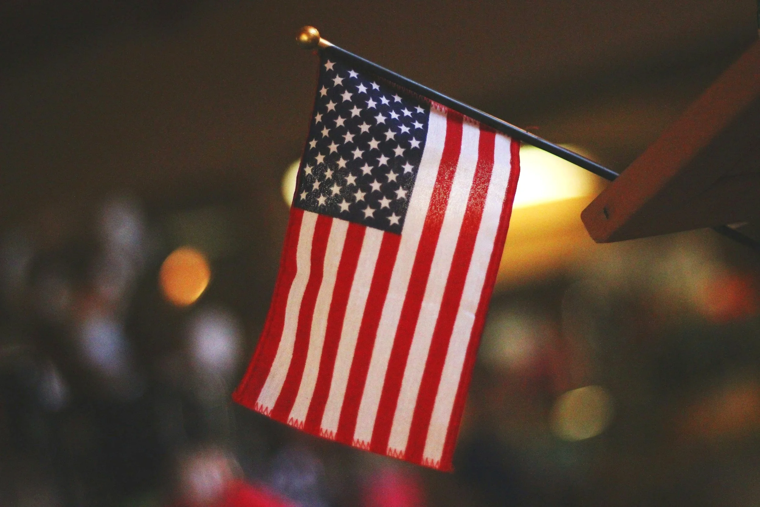 Small American flag hanging from a pole with a blurred background of lights. Bart Hebert for Judge