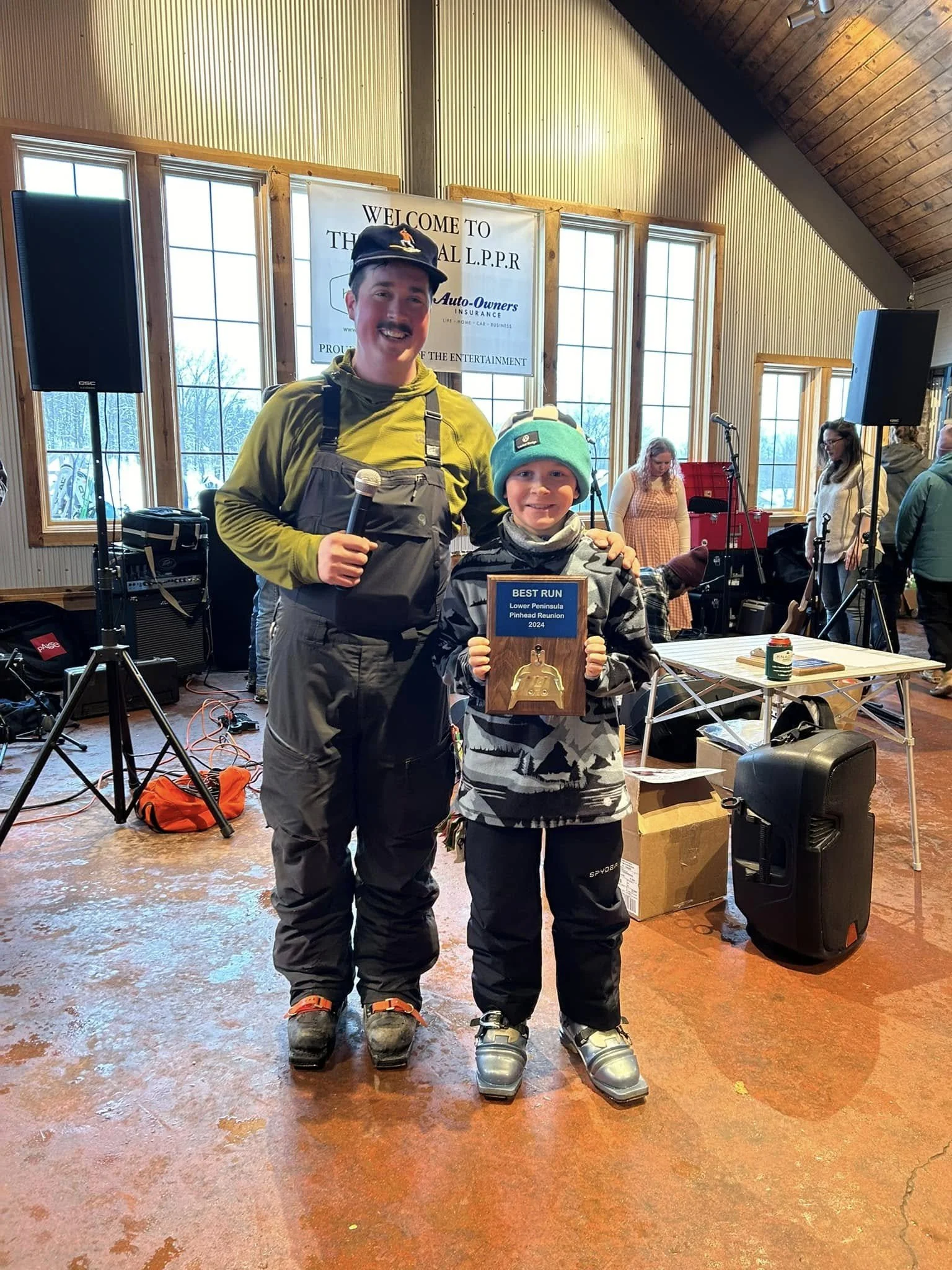 A boy and an adult man standing together indoors, with the boy holding a plaque that says 'Best Run Lower Peninsula Pinhead Reunion 2024.' They are smiling and dressed in winter clothing, with the man wearing ski or snowboarding gear and the boy wearing a knit hat and winter jacket. The background features large windows, a sign that reads 'Welcome to The Local LL.P.R. Auto-Owners Insurance,' and a few people and musical equipment.