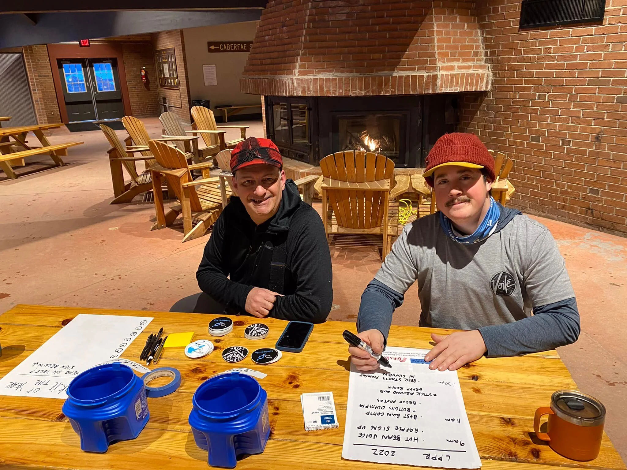 Two men sitting at a wooden table with writing materials, badges, and coffee mugs in front of a fireplace in a cozy indoor setting.