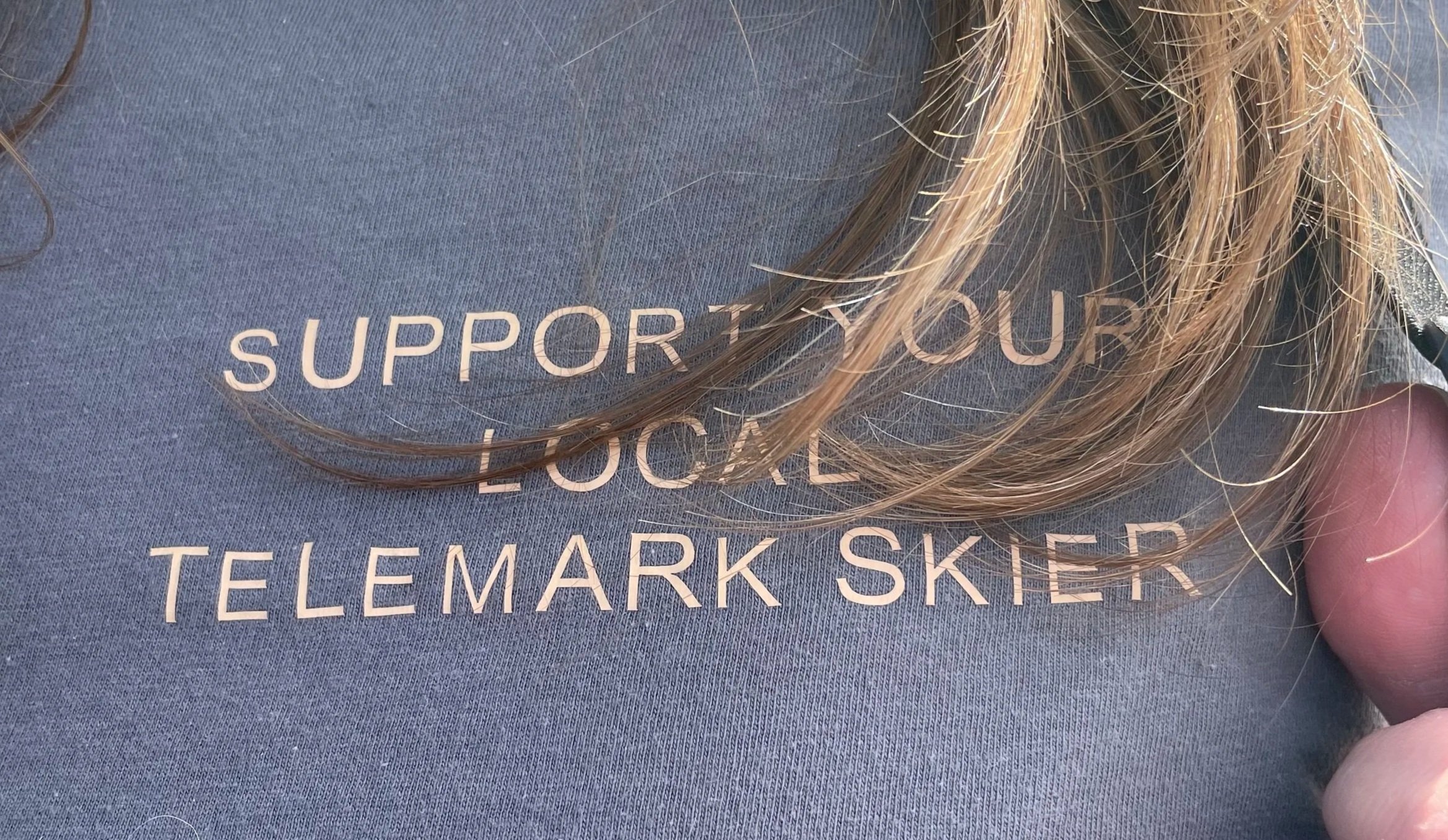 Close-up of a person's chest wearing a gray shirt with text supporting Local Telemark Skier, with blonde hair partially covering the words.