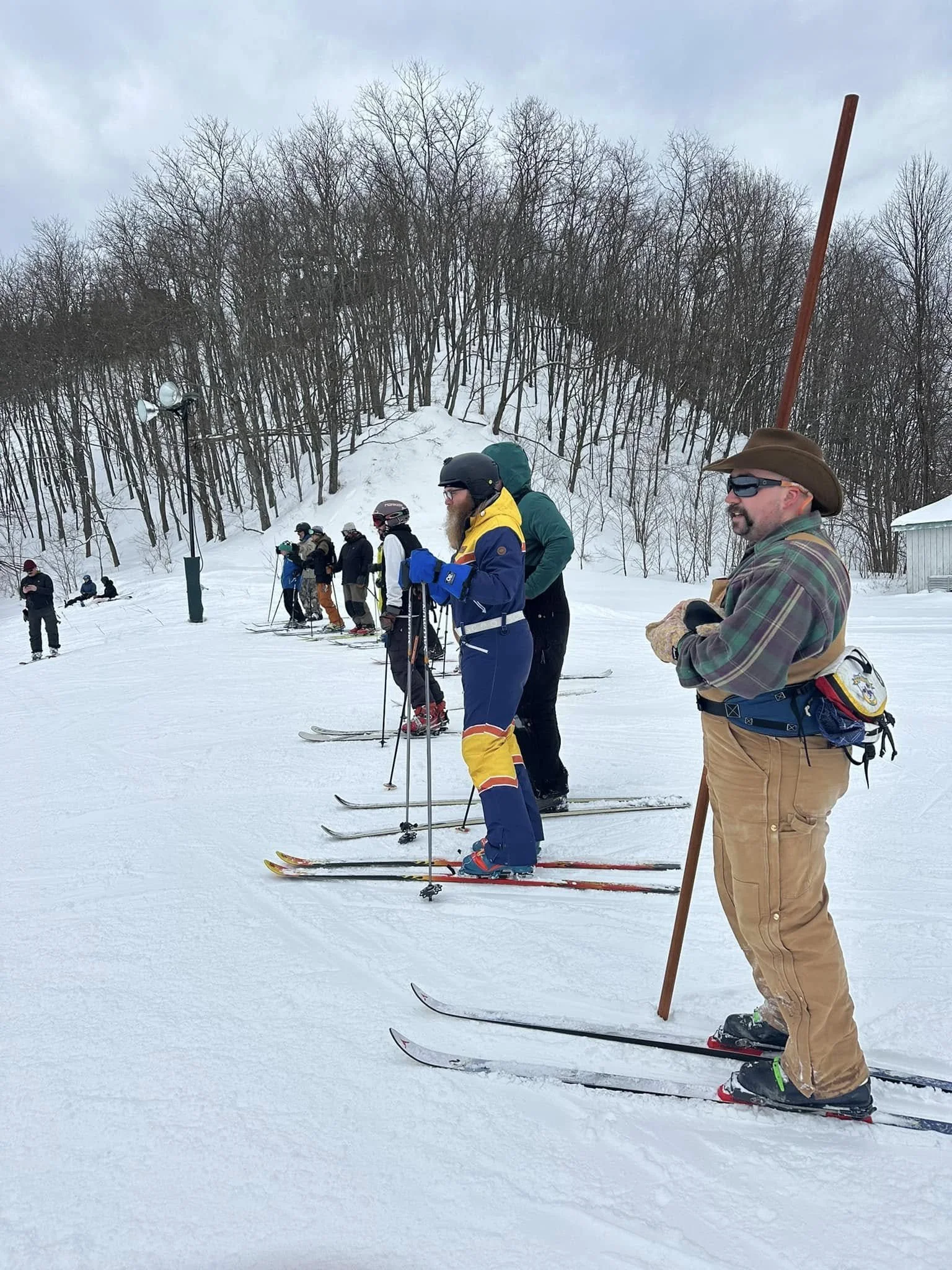 Group of people lined up in a row for cross-country skiing on a snow-covered landscape with leafless trees in the background.