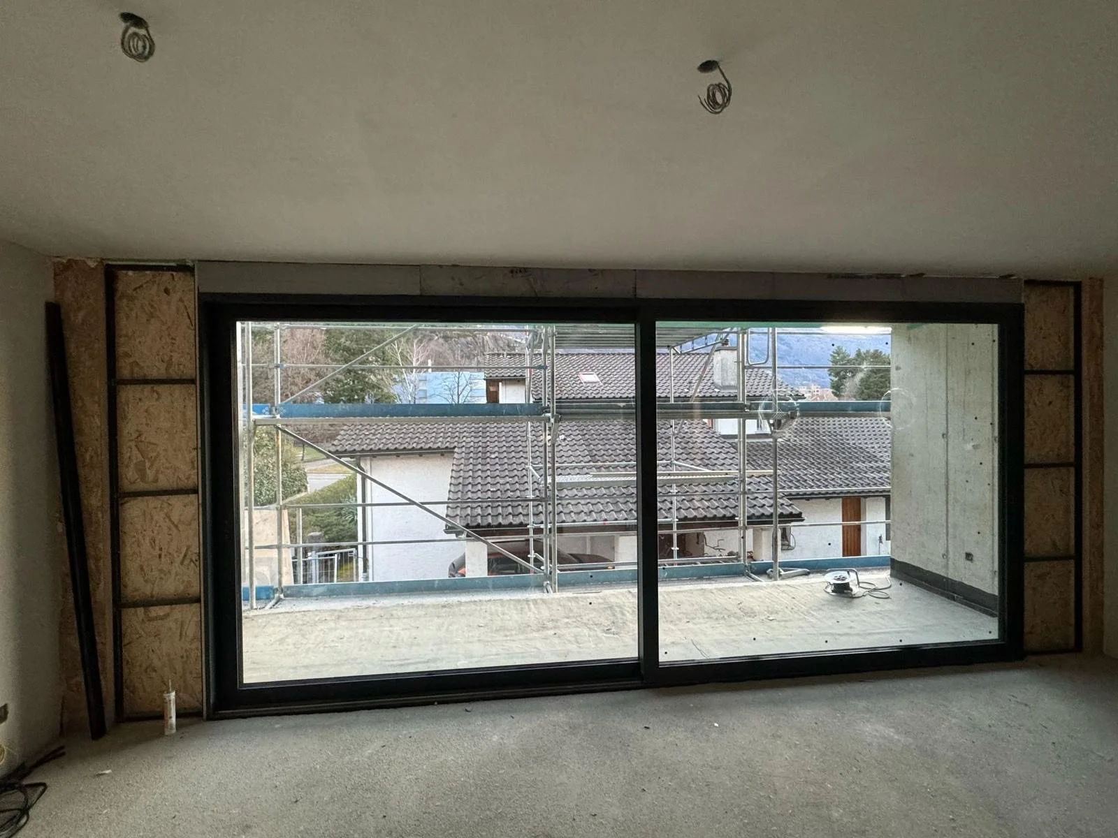 Interno di una stanza in costruzione con grande finestra in vetro e scaffalature laterali, vista sulle case e le montagne esterne.