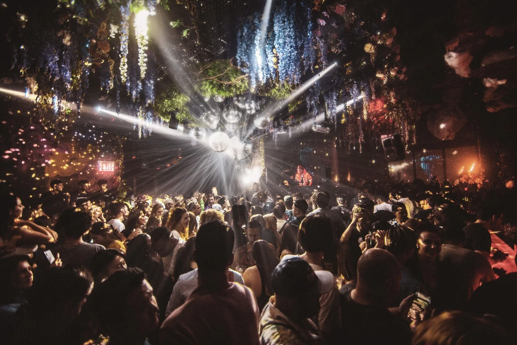 Crowd of people at a nightclub with colorful lights, hanging decorations, and a DJ performing on stage.
