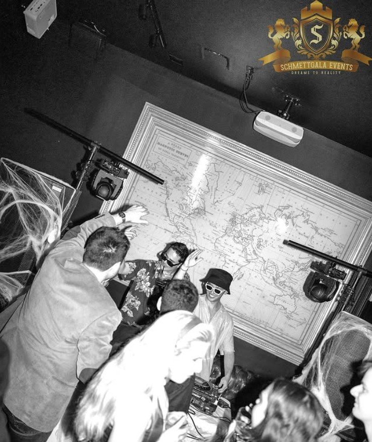 A DJ wearing sunglasses and a hat performs at a party with dancing guests, in a room decorated with cobwebs and a large world map on the wall.