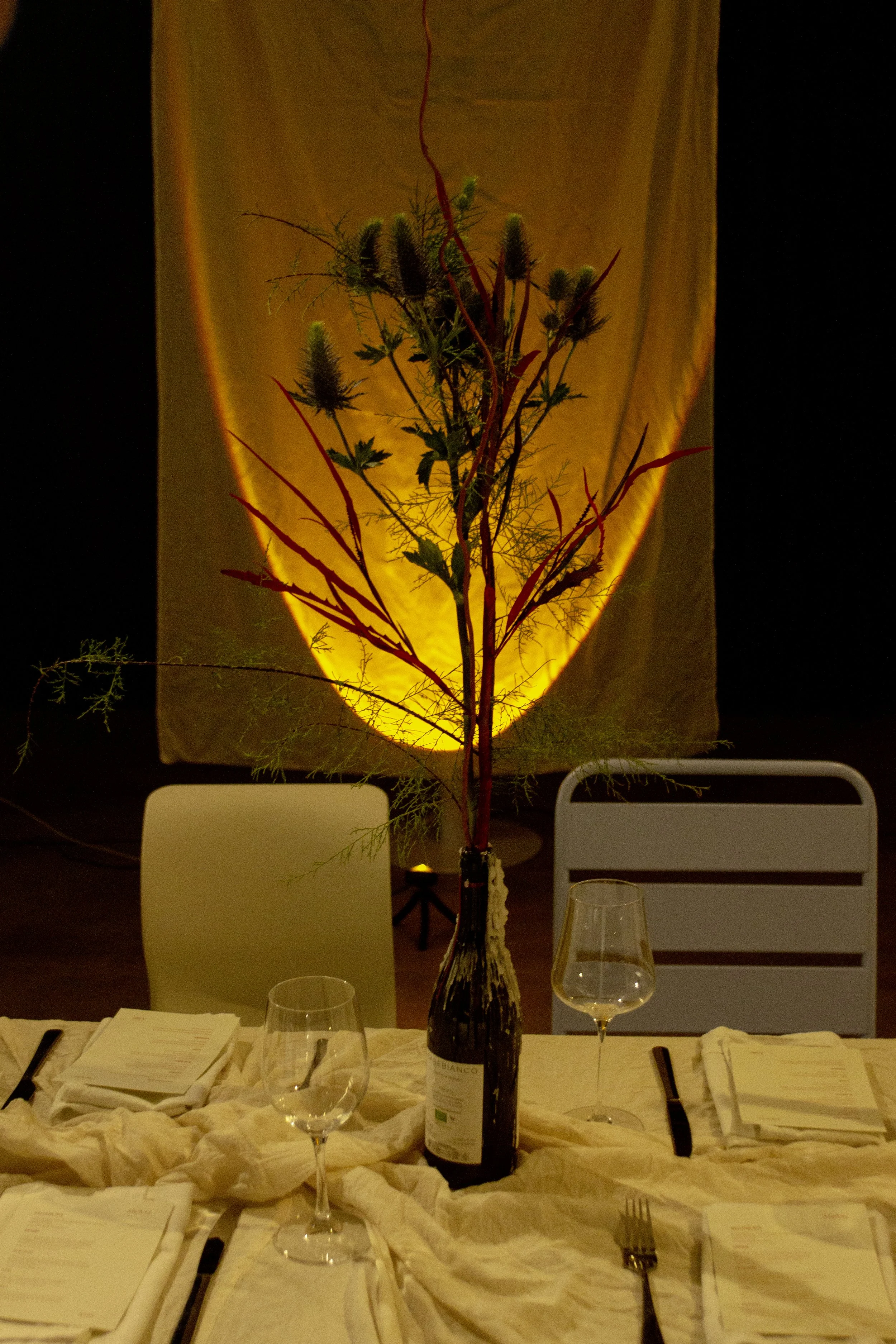 A dining table with chairs, set with menus, wine glasses, and black cutlery, with a centerpiece featuring a bottle with red twigs and green foliage, placed in front of a glowing yellow lamp