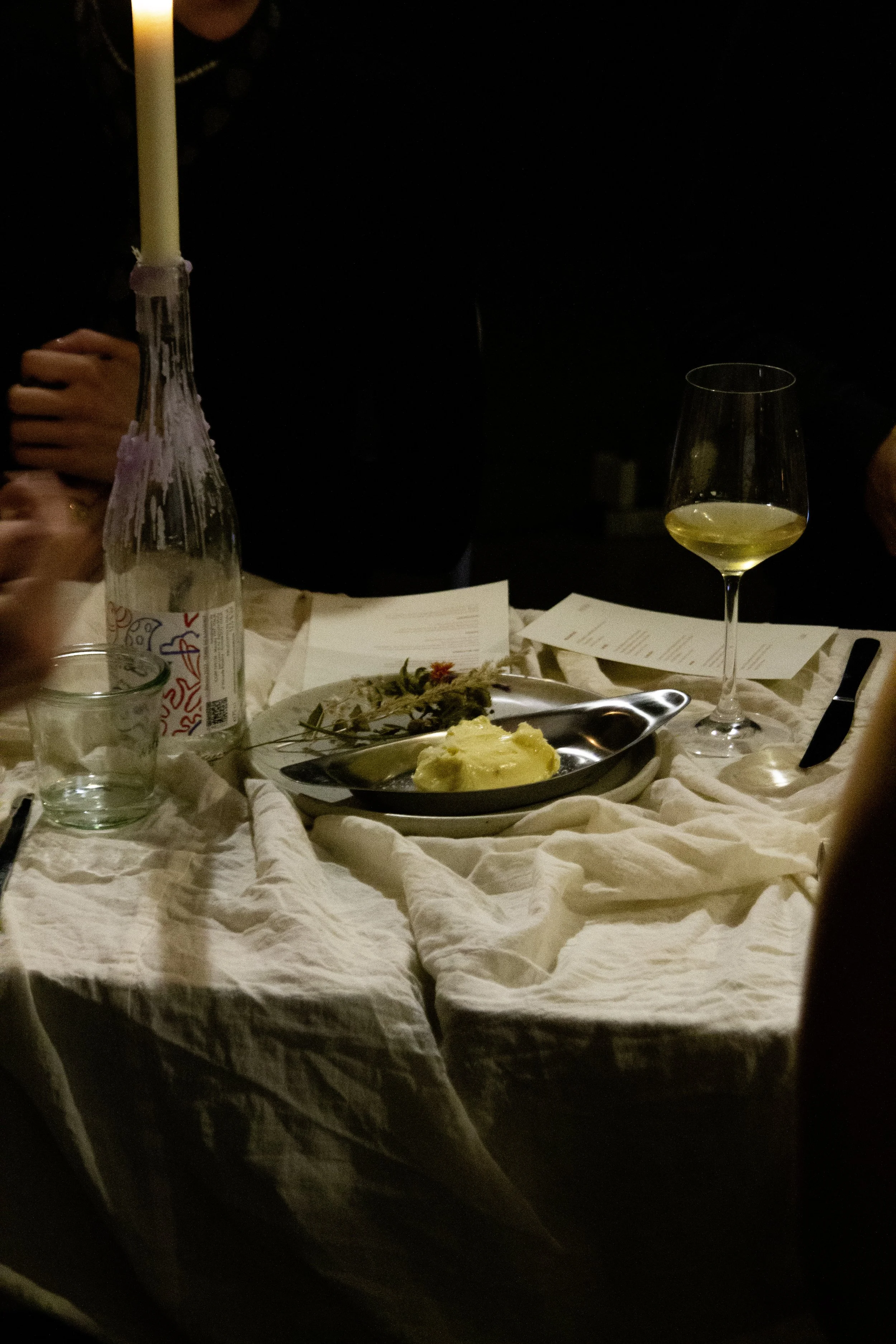 Table set with a candle, a bottle of sparkling water, a glass of white wine, a plate with mashed potatoes, and some papers, with people sitting around.
