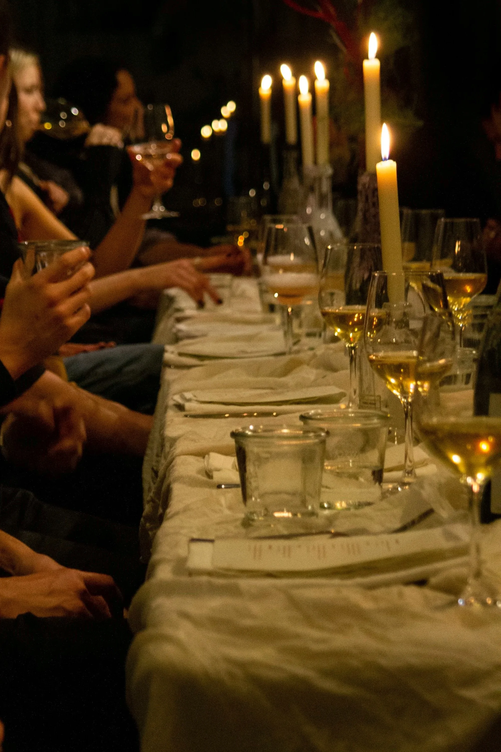 People sitting at a dinner table with lit candles and glasses of white wine, enjoying a celebration or gathering in a dimly lit setting.