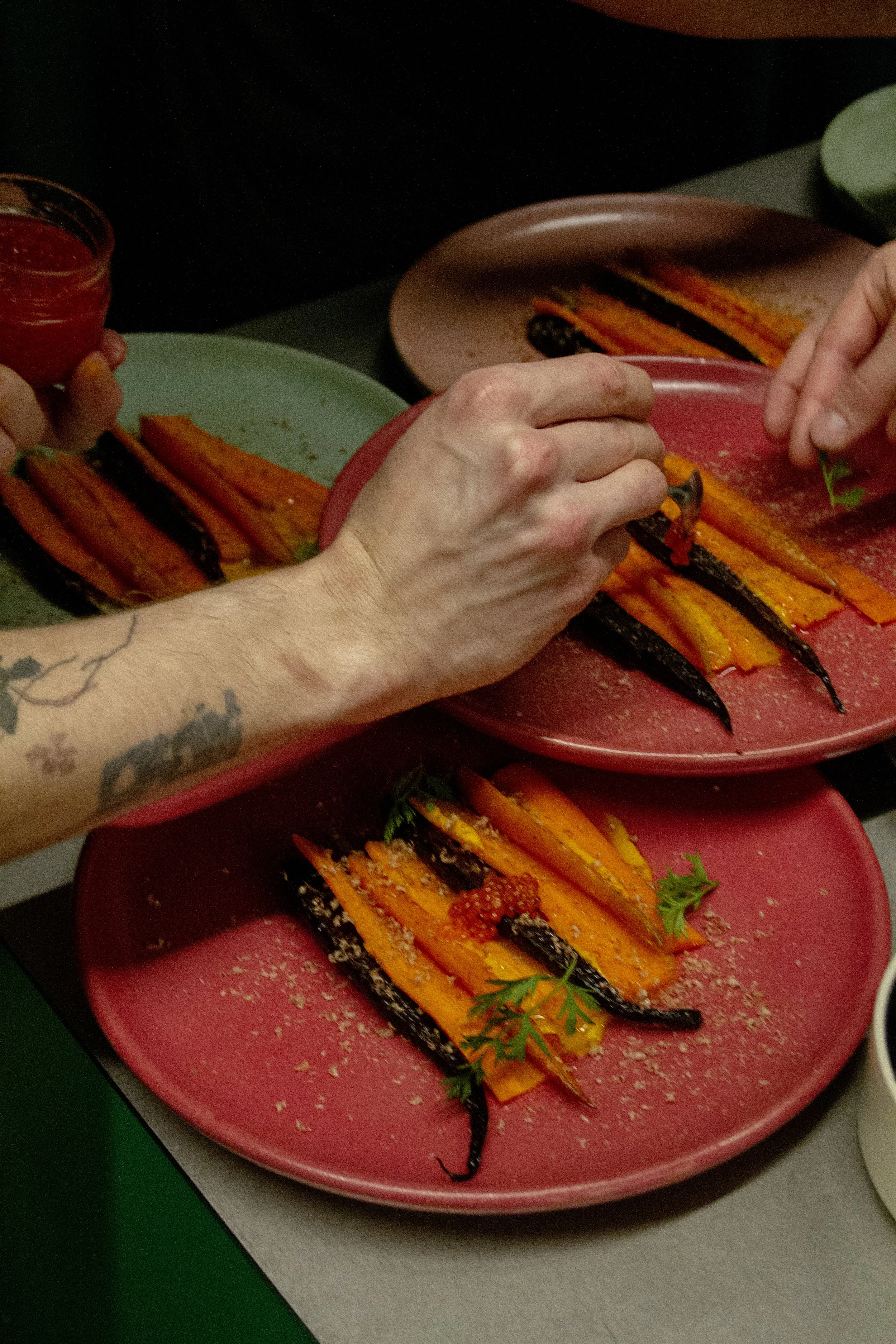 Person with tattooed arm preparing roasted carrots on pink and green plates in a kitchen.
