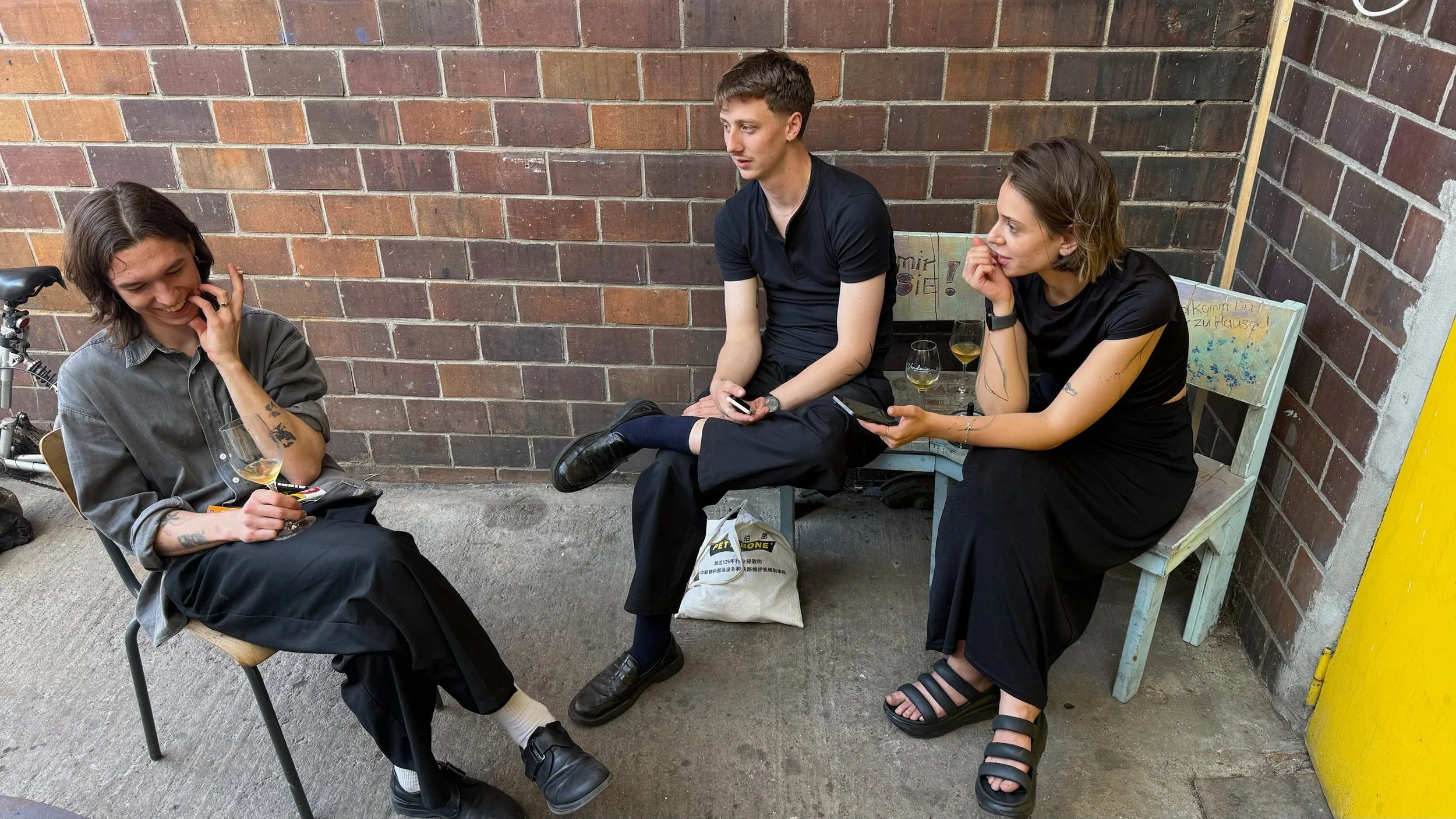 Three young adults sitting outdoors against a brick wall, engaging in conversation. One of them is holding a wine glass, and there are two glasses of wine on a bench beside them.
