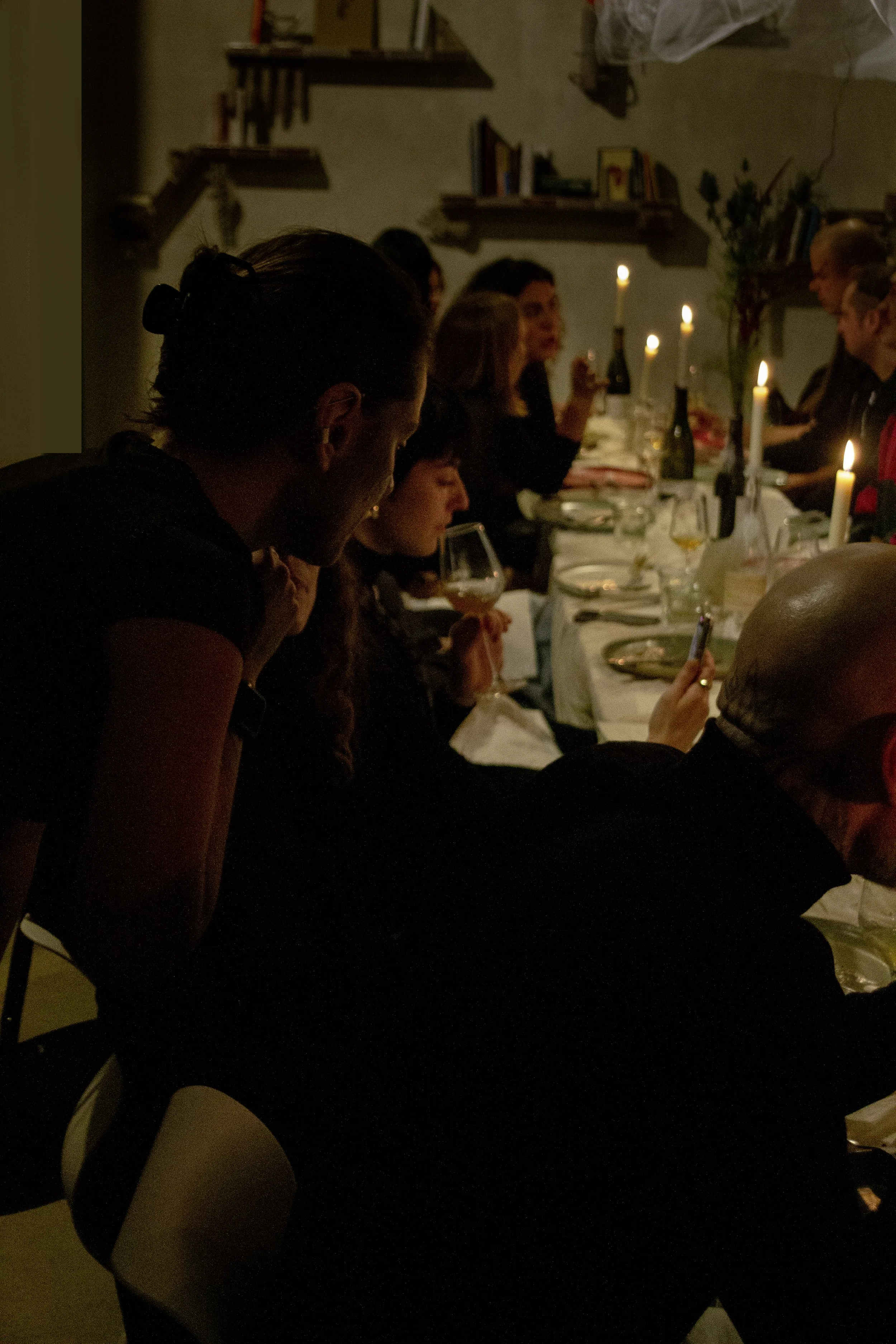 People gathered around a table with lit candles, dinnerware, and wine glasses in a cozy, dimly lit setting.