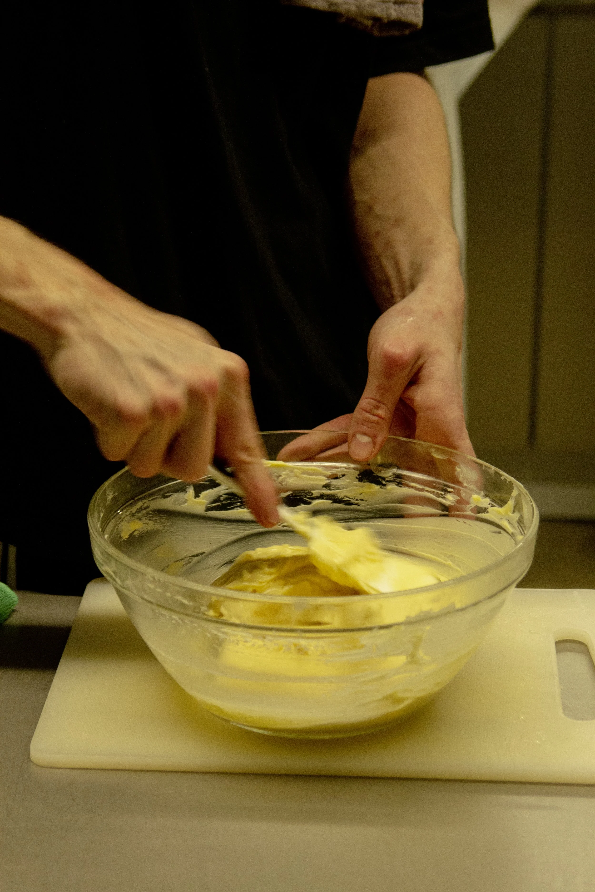 A person mixing yellow batter in a clear glass bowl on a white cutting board, using a spoon, in a kitchen setting.
