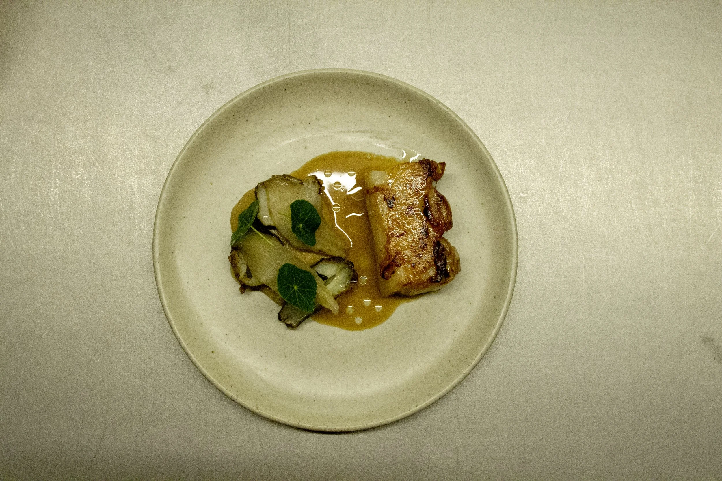 A plated gourmet dish featuring a piece of cooked meat, likely chicken or fish, accompanied by roasted vegetables and garnished with green leaves, served on a beige ceramic plate.