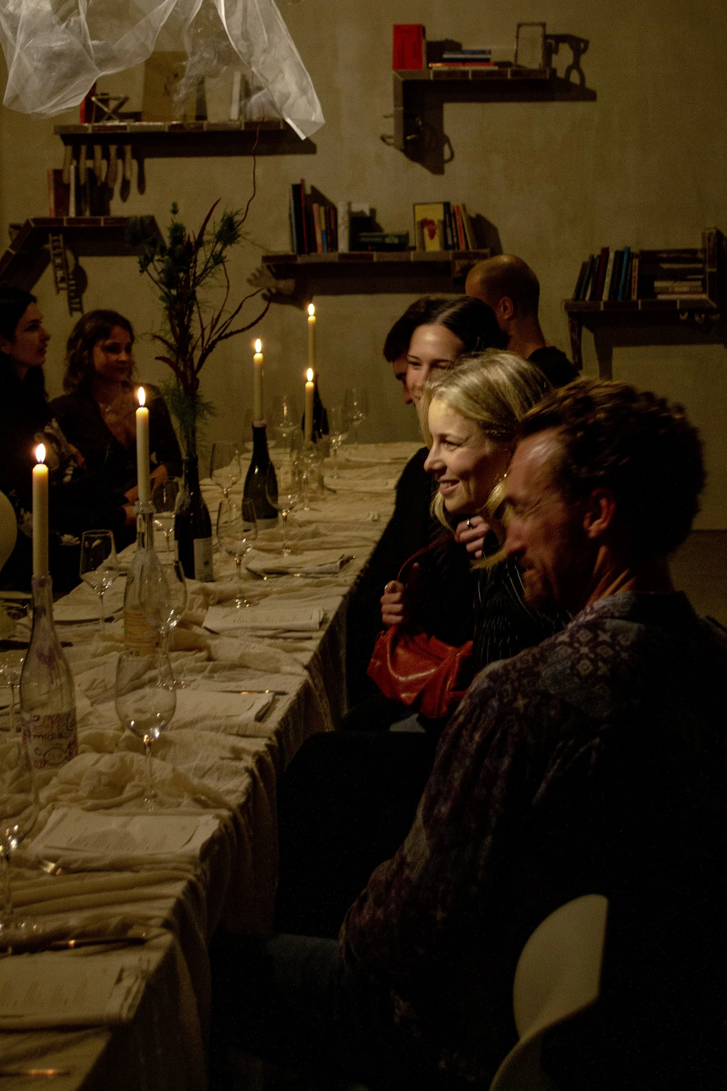 People sitting at a long dinner table with lit candles in a dimly lit room, enjoying a social gathering.