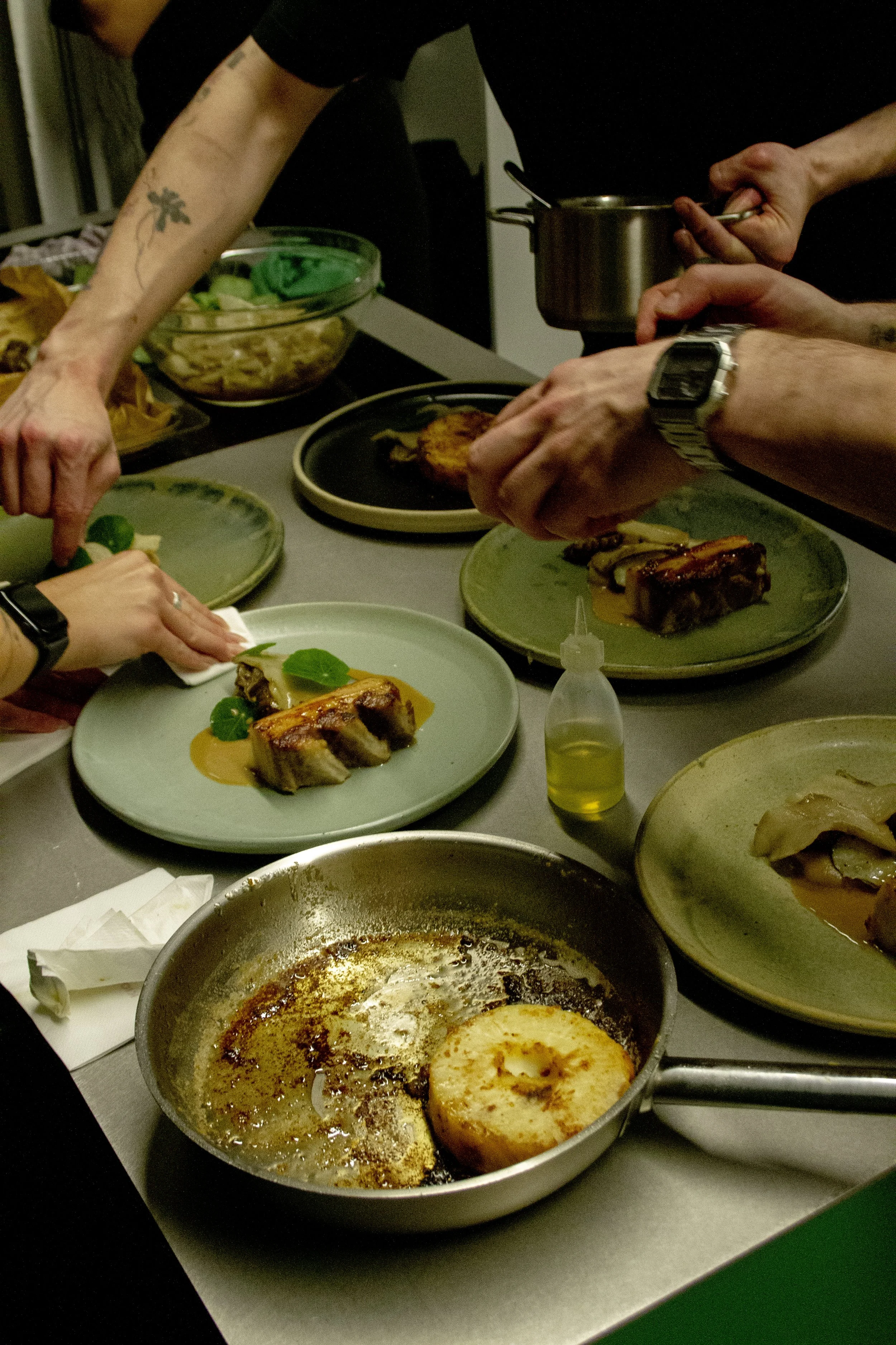 People plating and serving food, including pork belly with sauce, at a dinner event with dishes, sauces, and kitchen items on the table.