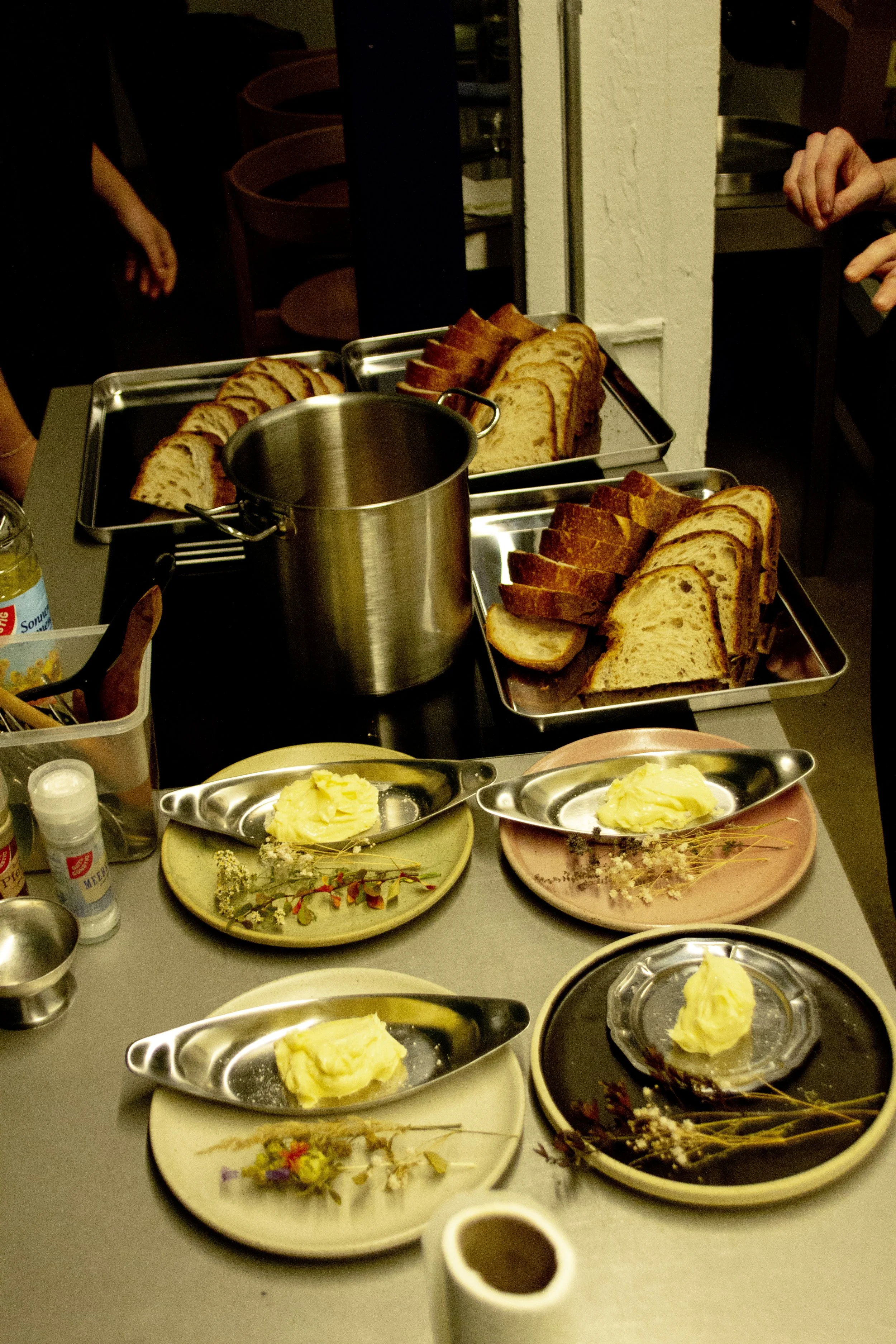 Arrangement of sliced bread, butter, and garnishes on plates and trays on a table, with a pot and condiments visible, in a dining setting.