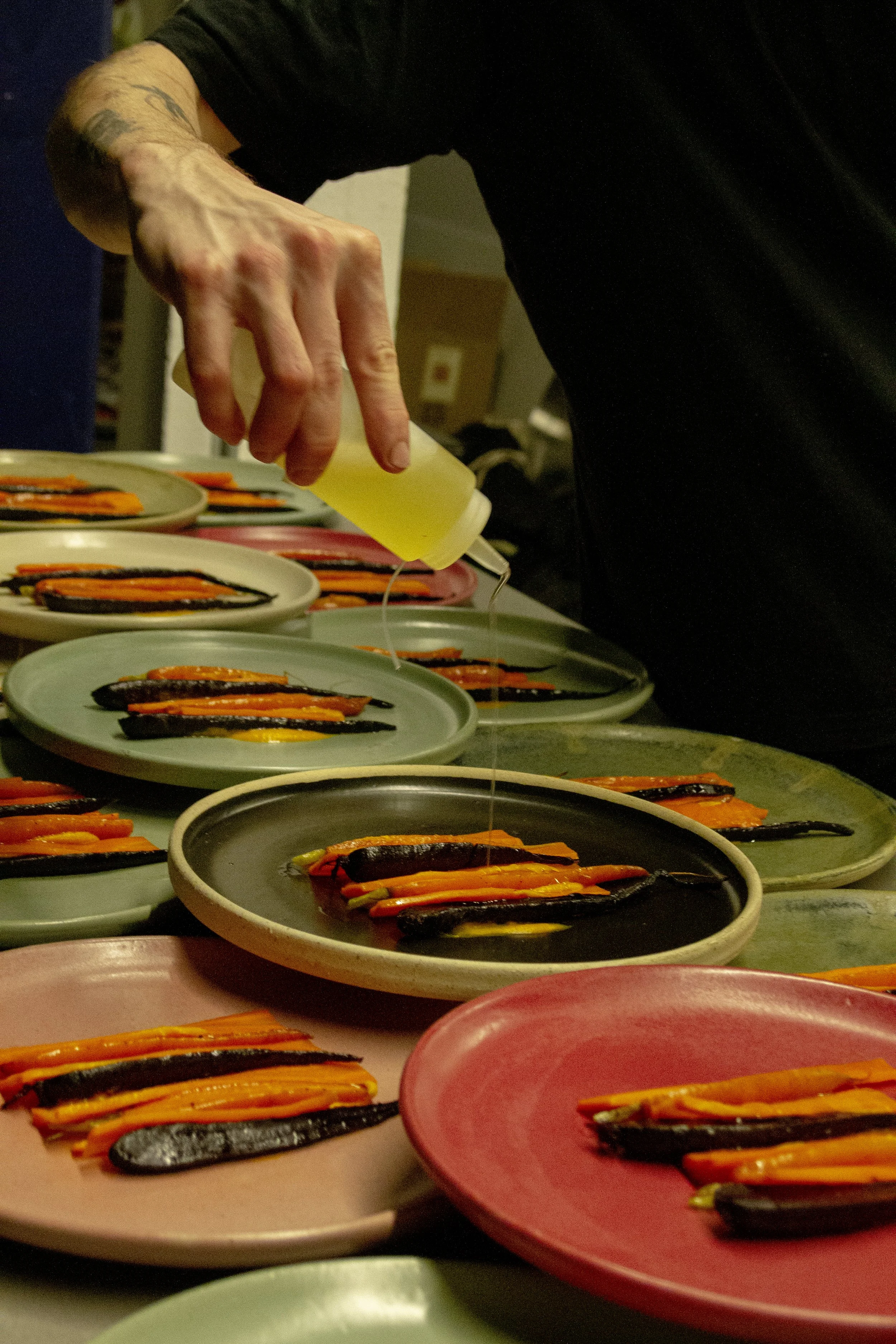 A person drizzles oil onto grilled vegetables on colorful plates, likely preparing dishes in a kitchen or restaurant. The vegetables appear to be carrots and possibly eggplant.
