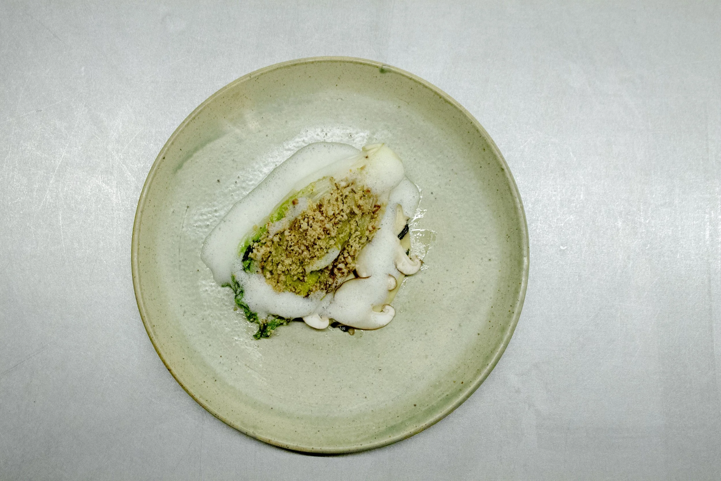 A small dish with a stuffed vegetable topped with foam and crumbs, served on a beige plate.