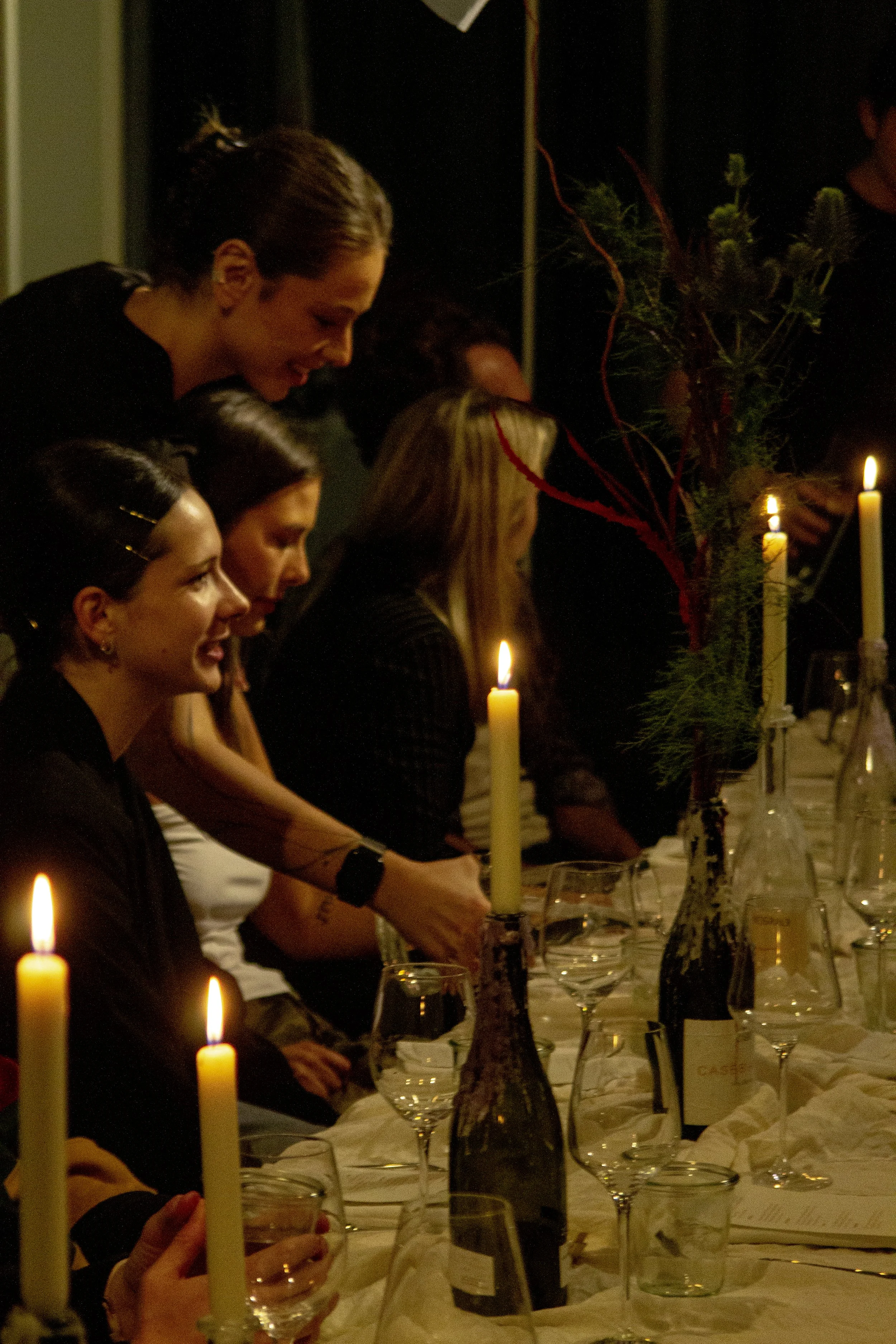 A group of women sitting at a dining table decorated with lit candles and vases with floral arrangements, during a dinner event.