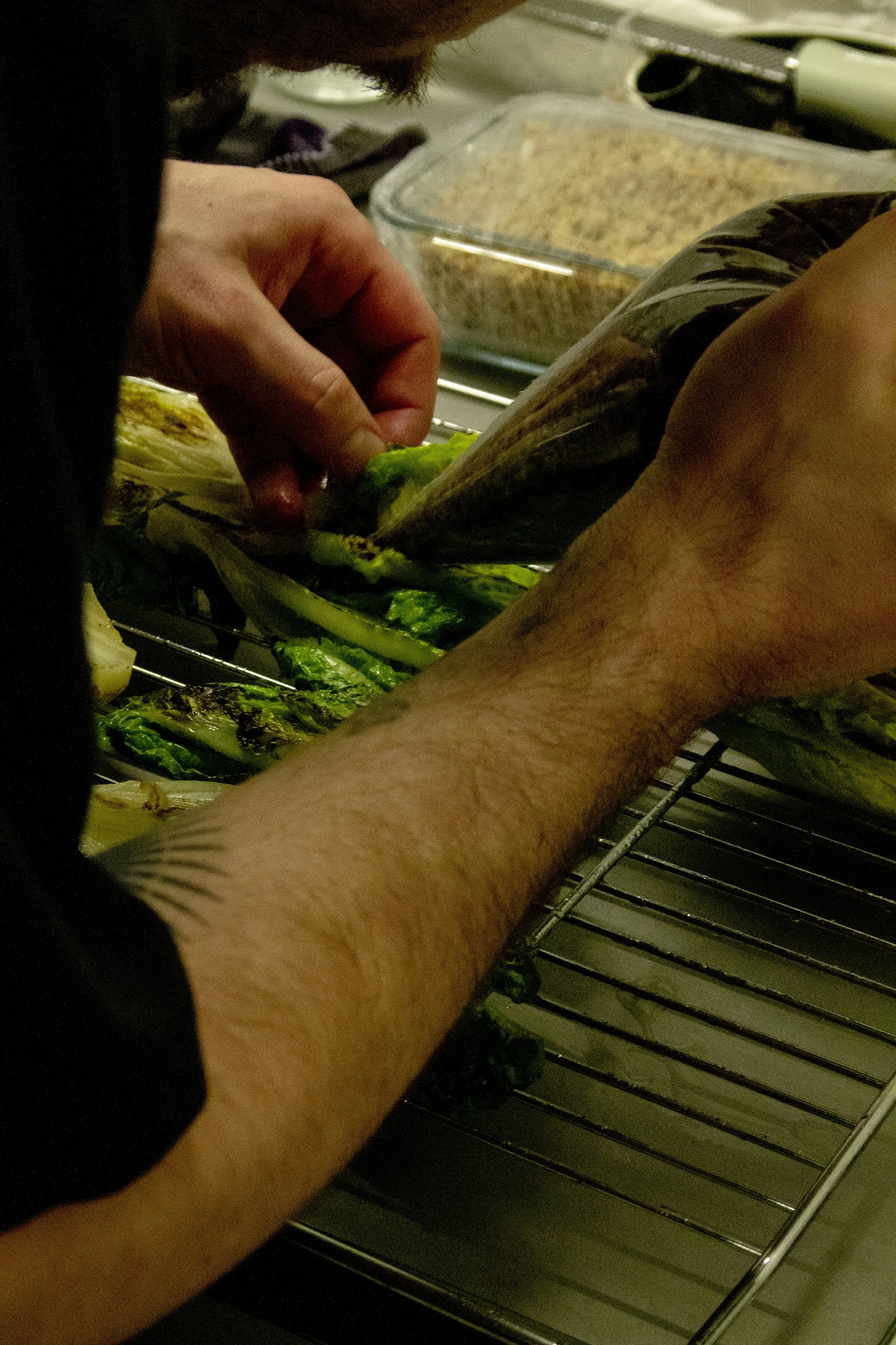 Close-up of a person grilling zucchini on a barbecue grill with a hand holding one piece and a container of breaded food in the background.