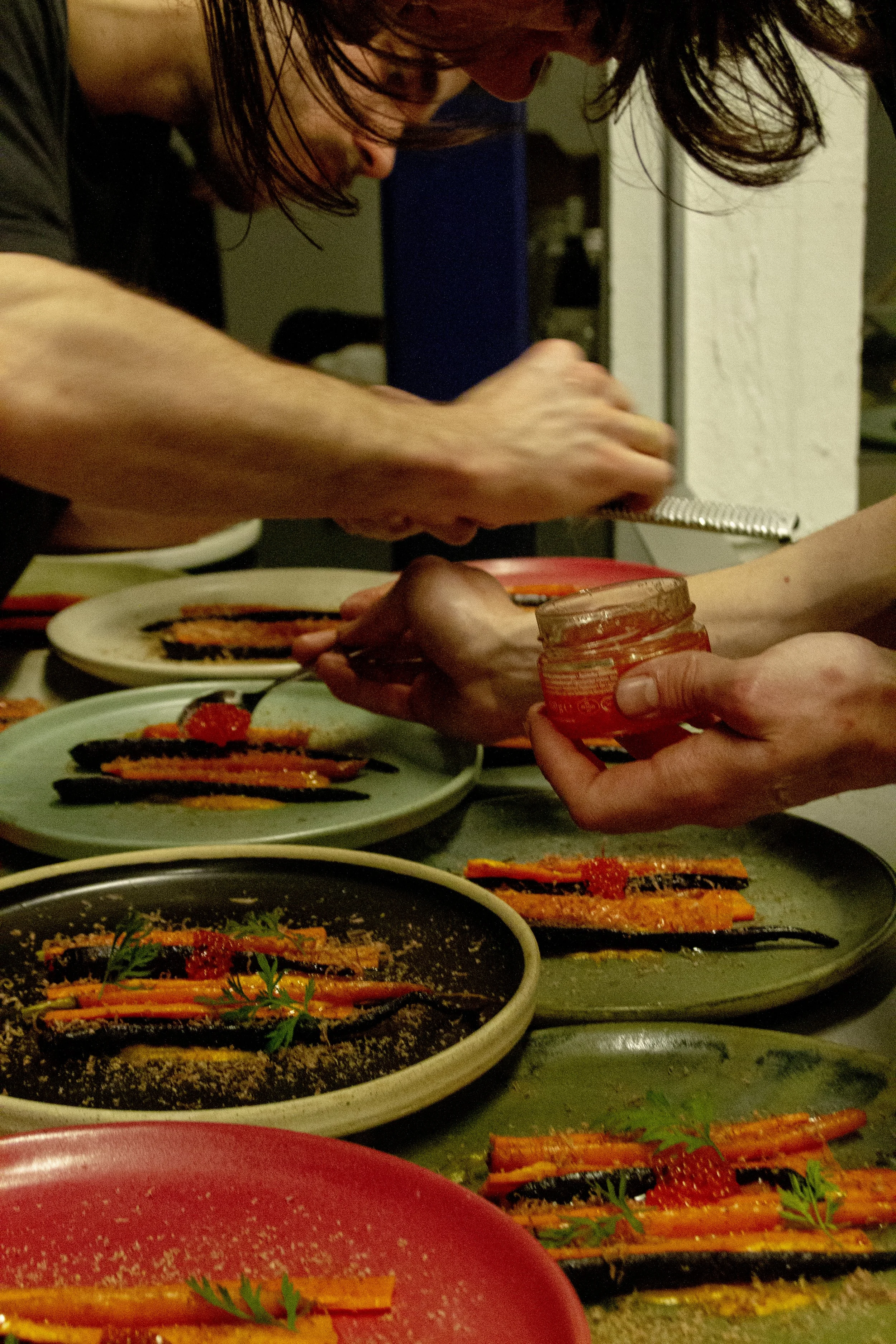 Two people preparing and garnishing plated dishes with carrots and strawberries in a kitchen.