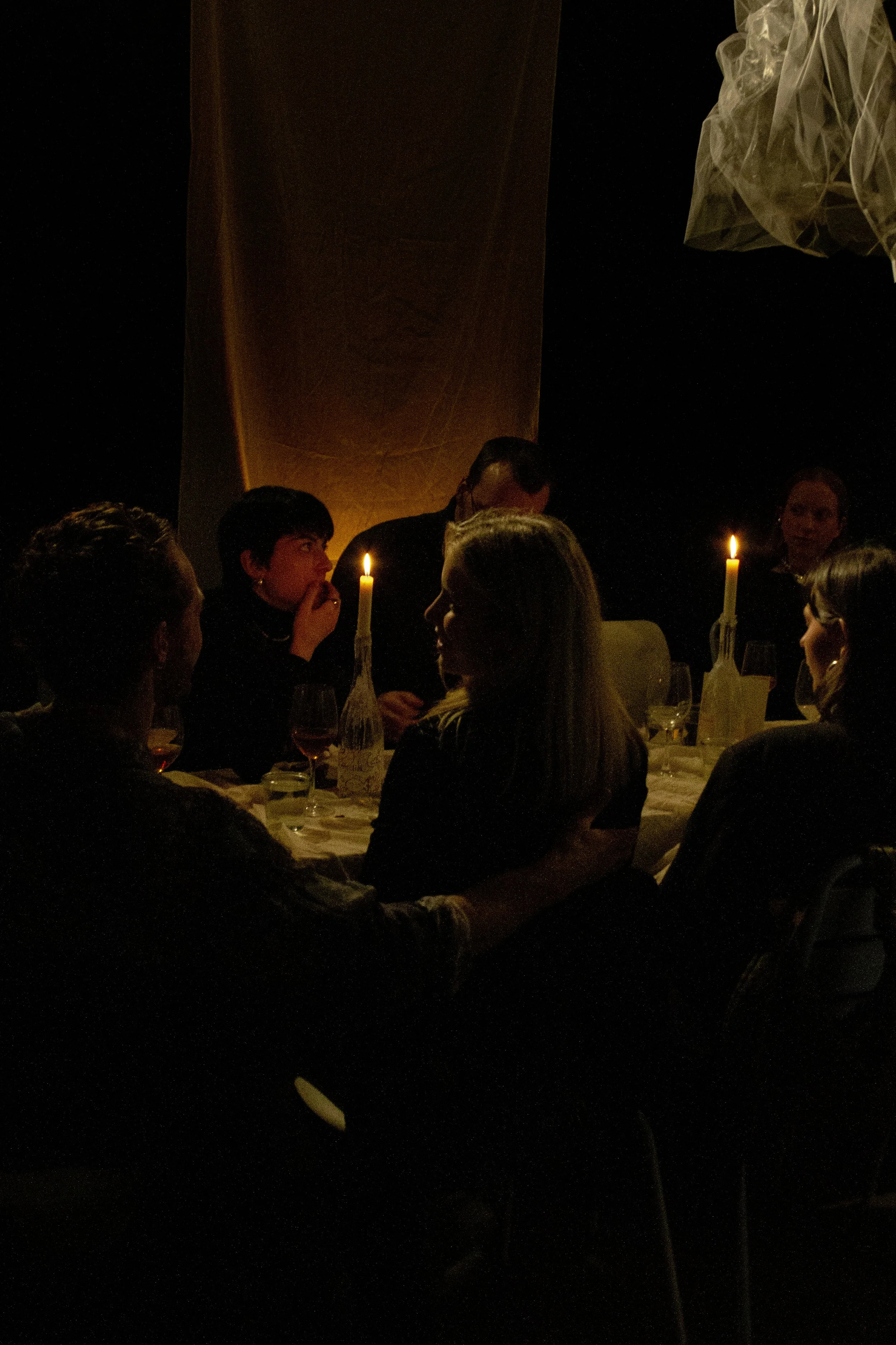 Group of people sitting around a dinner table with candles in a dimly lit room, engaged in conversation.