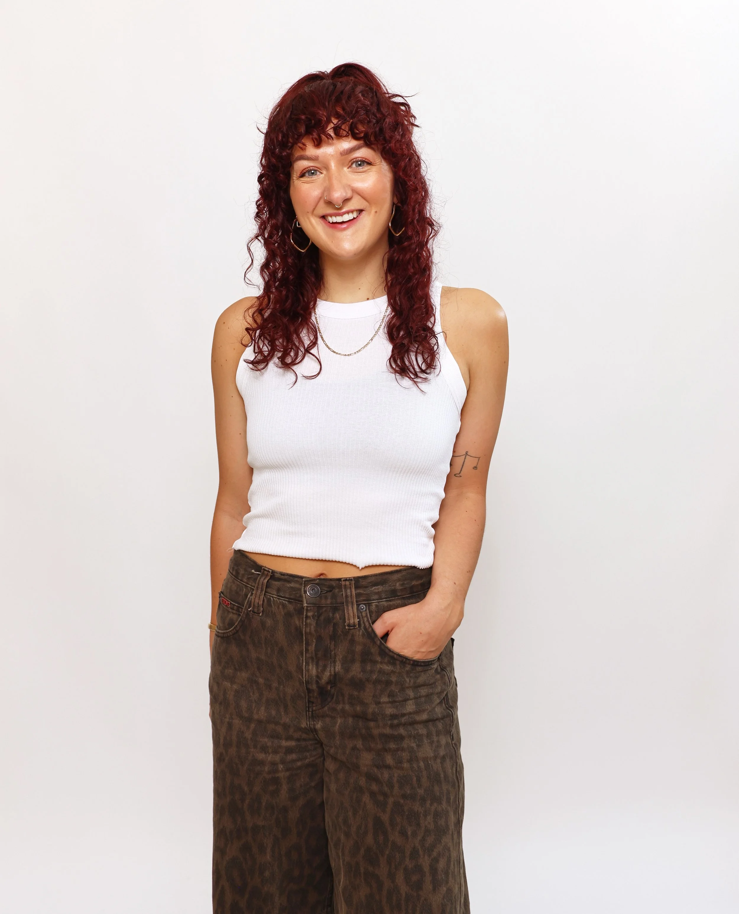 A young woman with curly red hair, wearing a white sleeveless top and brown patterned pants, standing against a plain white background, smiling at the camera.