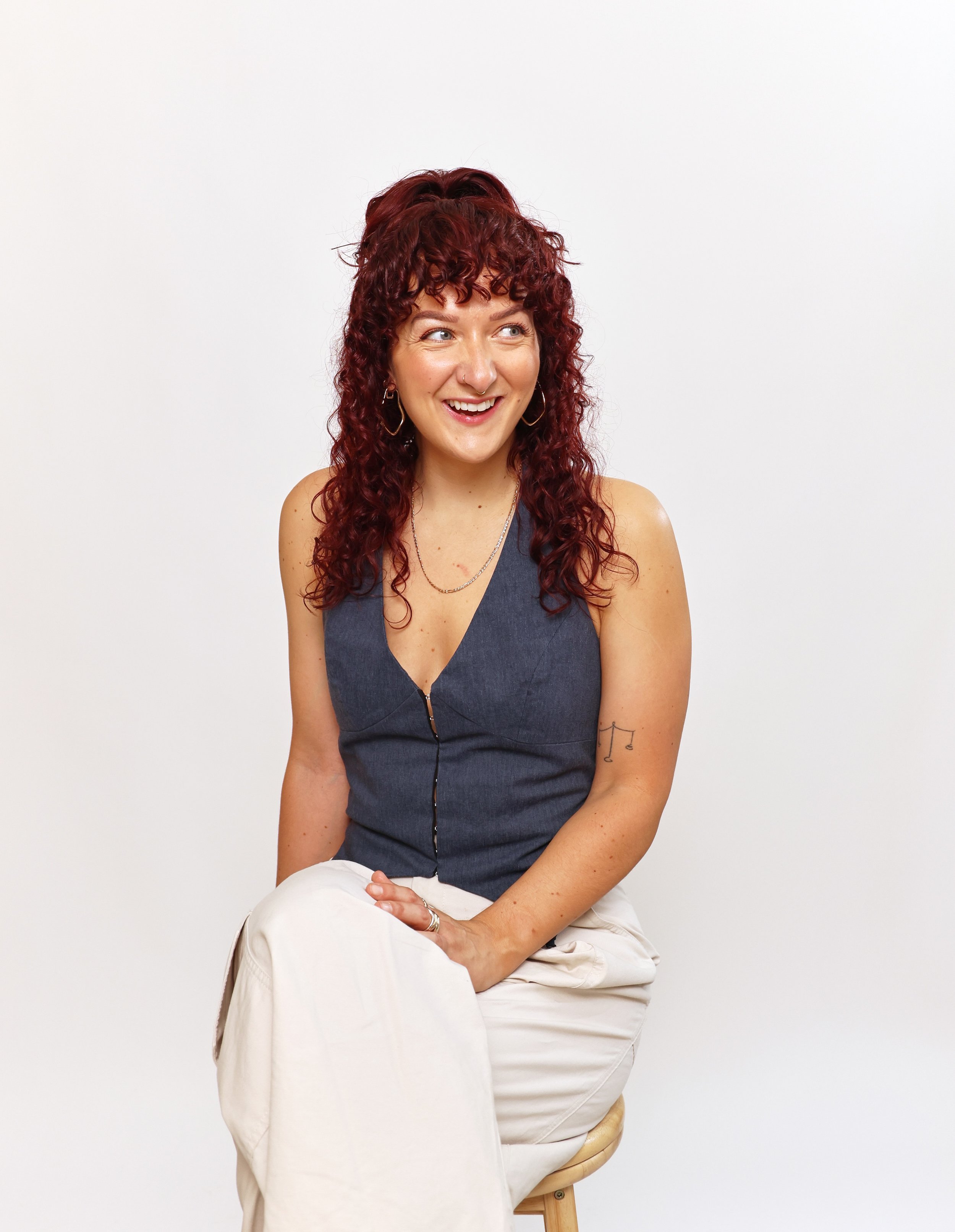 A woman with curly red hair, wearing a sleeveless denim top and white pants, sitting on a wooden stool against a plain white background, smiling and looking to her right.
