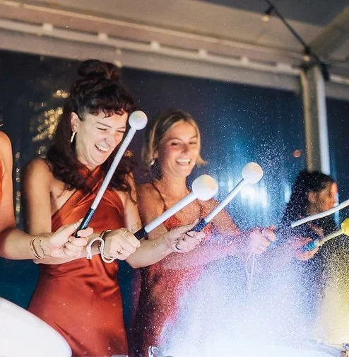 Three women smiling and playing with colorful light sticks at a celebration or party under a canopy.