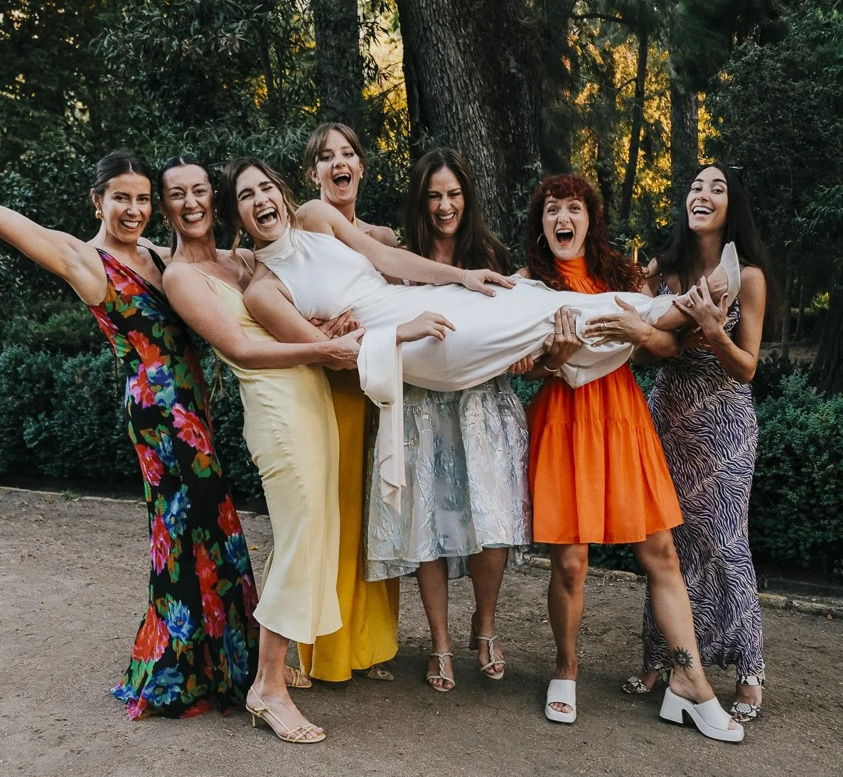 Group of women outdoors, lifting a woman in a white dress, all smiling and laughing, with lush green trees and sunlight in the background.