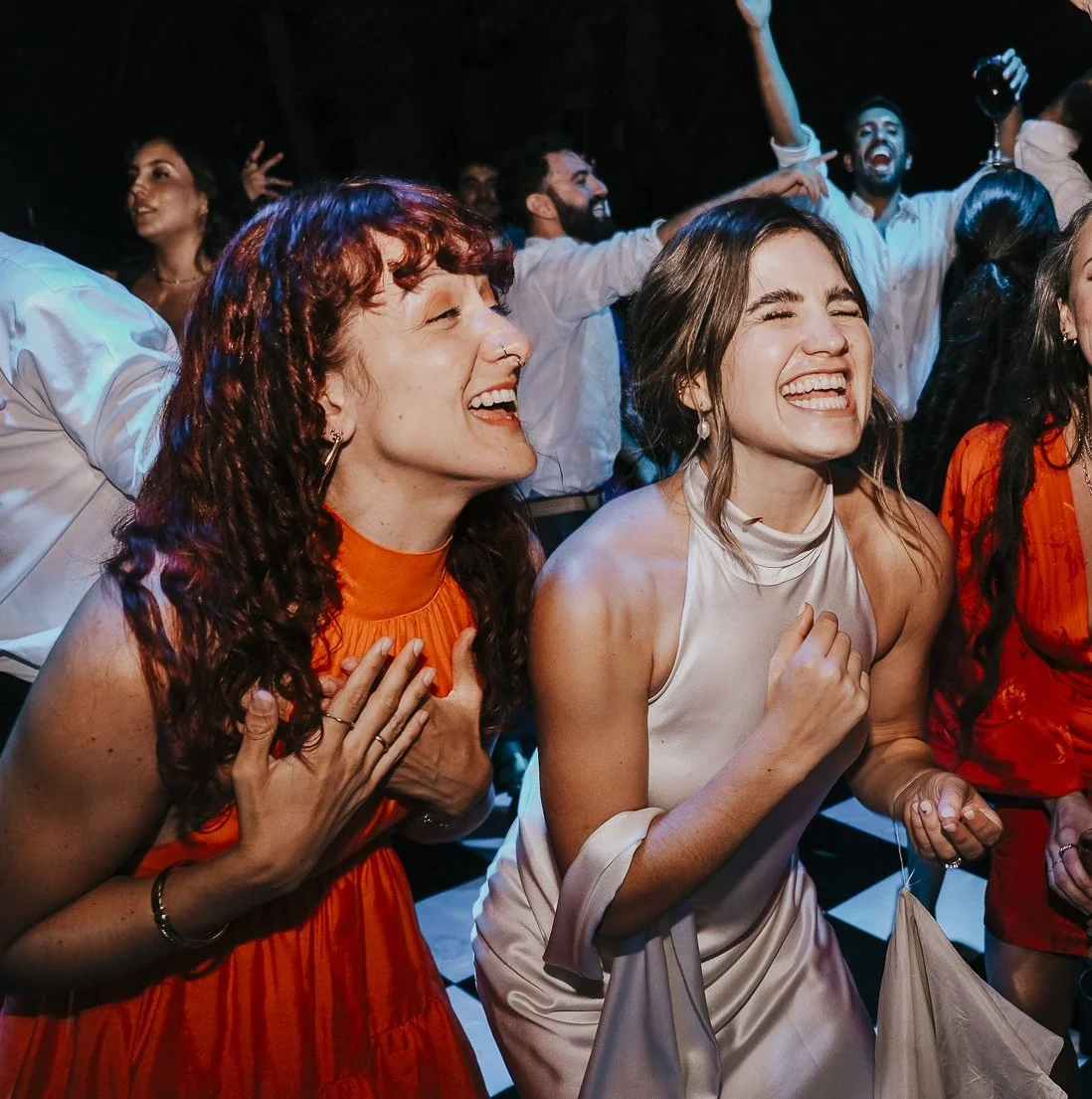 People at a lively party or celebration, laughing and dancing, with some holding drinks and others with hands on their chests, wearing colorful outfits.