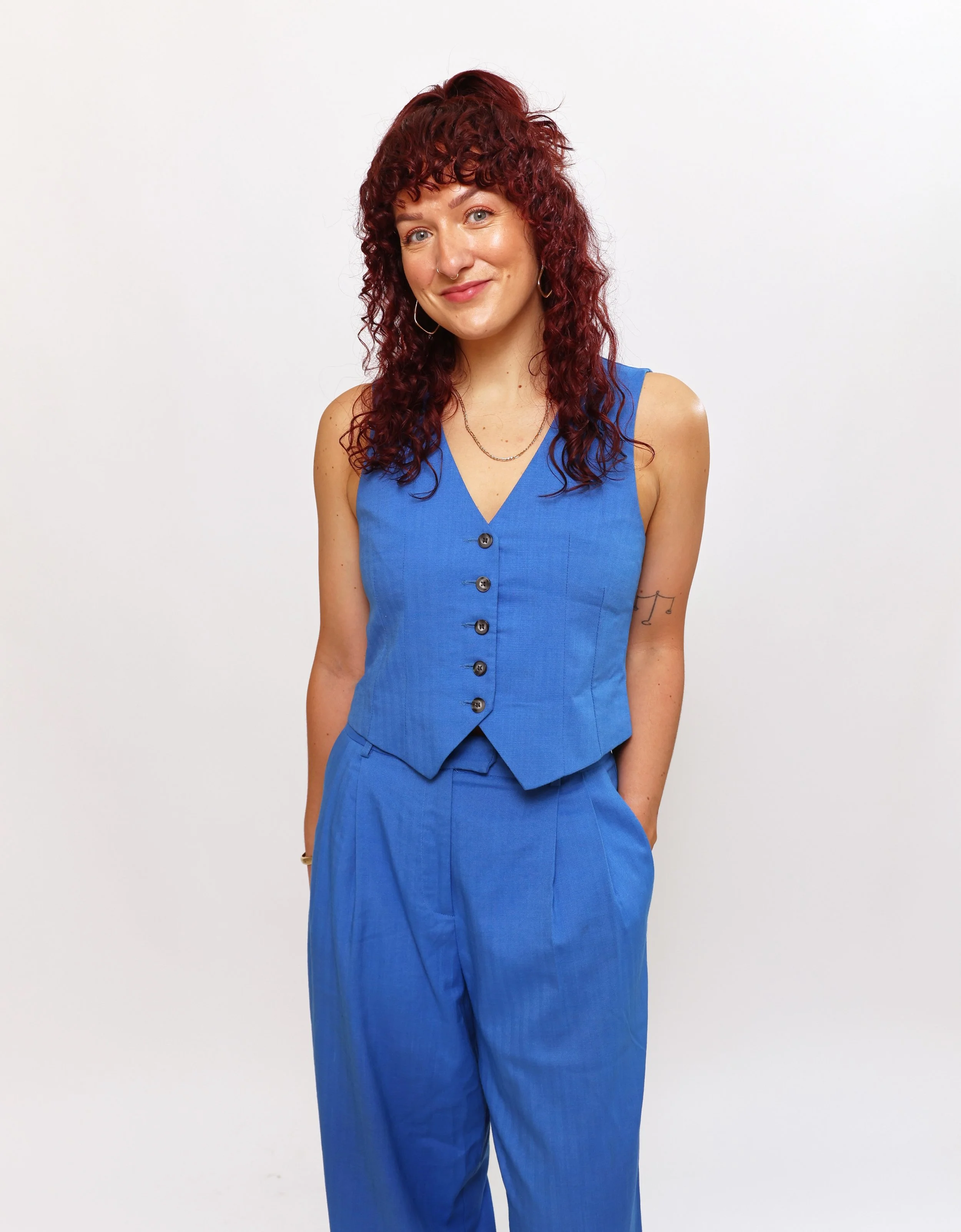A woman with curly red hair smiling at the camera, wearing a sleeveless blue vest and matching blue pants, standing in front of a white background.