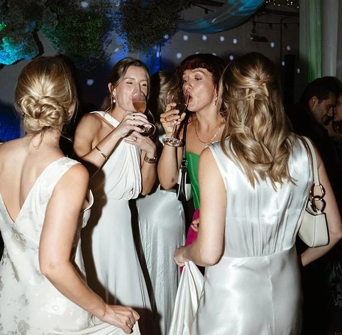 Four women in elegant dresses drinking wine at a social event or party with dim lighting, decorations, and other guests in the background.
