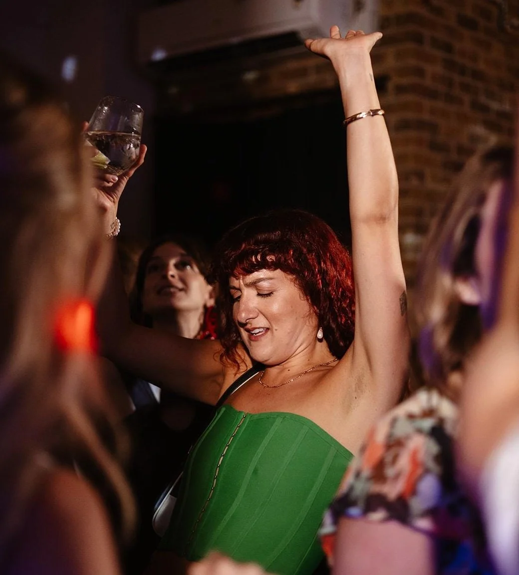 A woman with short curly red hair dancing with her eyes closed in a lively party environment, wearing a green strapless dress and jewelry, with other people around her holding drinks and socializing.