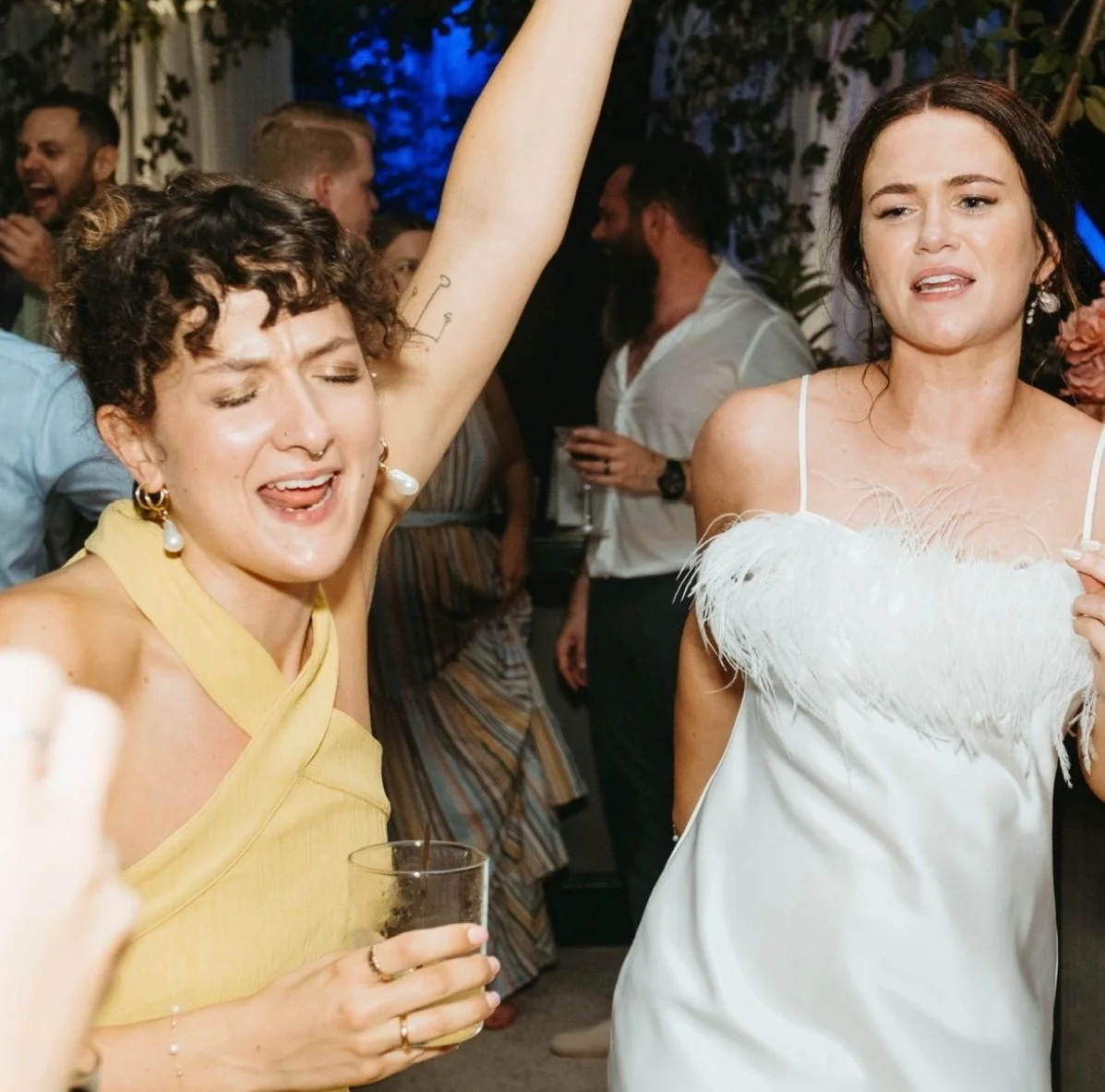 Two women dancing and enjoying a party, one with curly hair, earrings, and a nose ring, wearing a yellow top, and the other with long dark hair, wearing a white dress with feather details, at a lively social gathering.