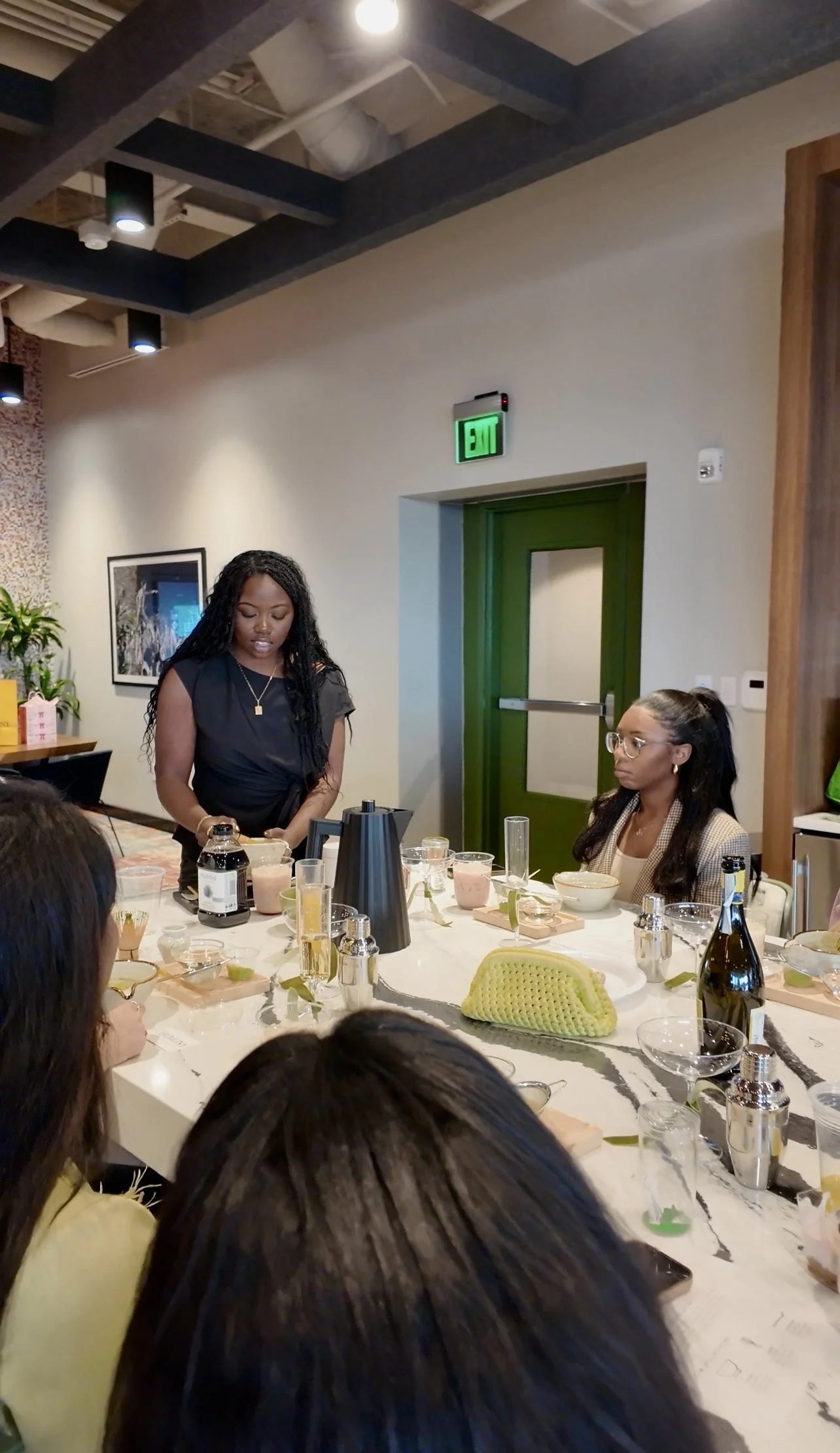 A group of women seated around a table during a tasting or presentation, with one woman standing and speaking, in a modern indoor setting with artwork and a plant in the background.