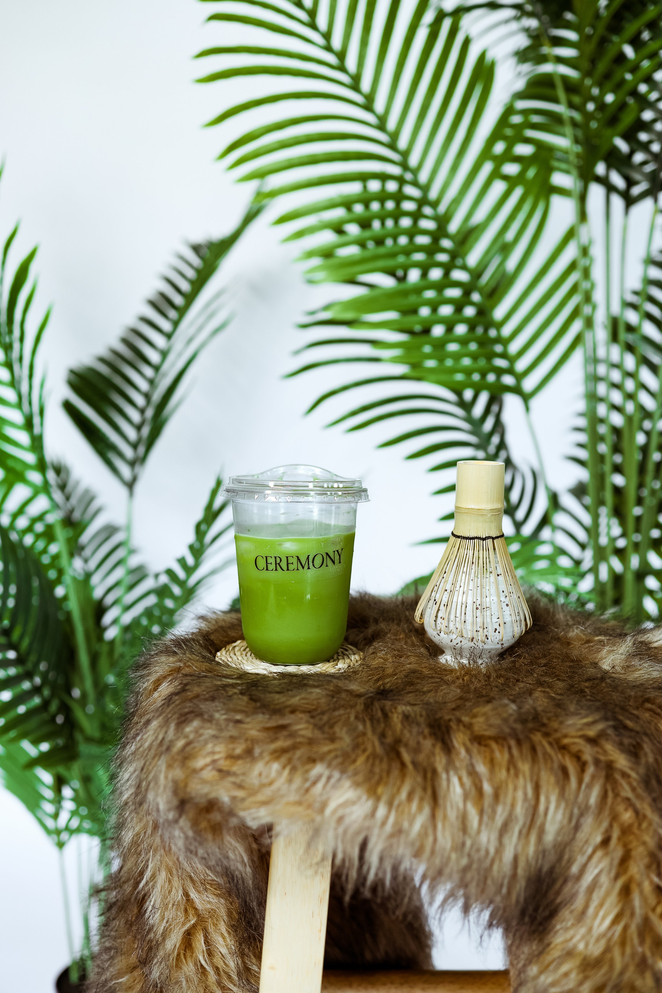 A small table covered with a furry brown cloth, holding a green drink labeled 'CEREMONY' in a clear plastic cup with a lid, and a decorative white and beige vase, in front of large green palm leaves.