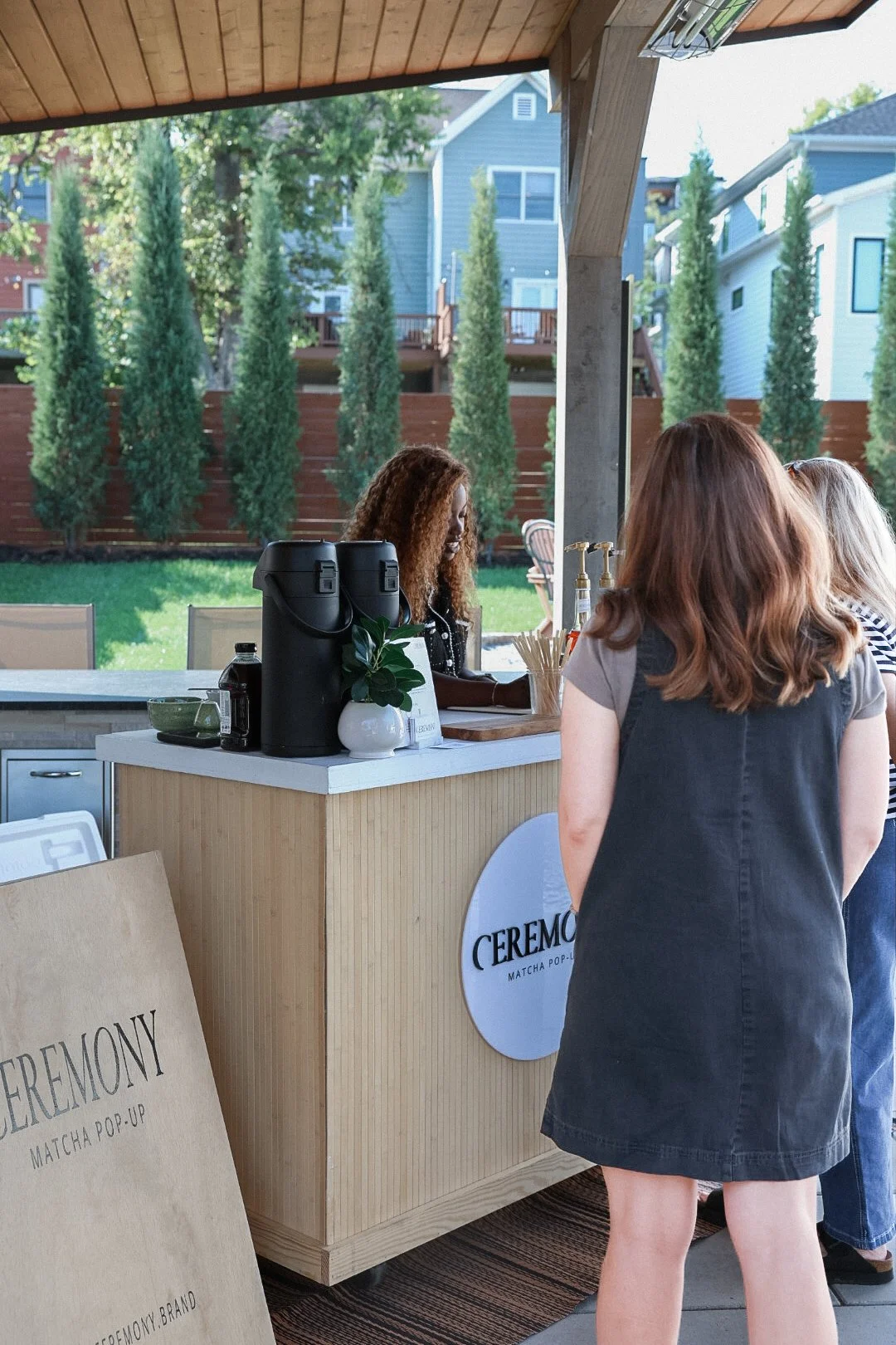 People standing at a matcha bubble tea pop-up counter indoors with a view of a backyard with tall trees and houses outside.