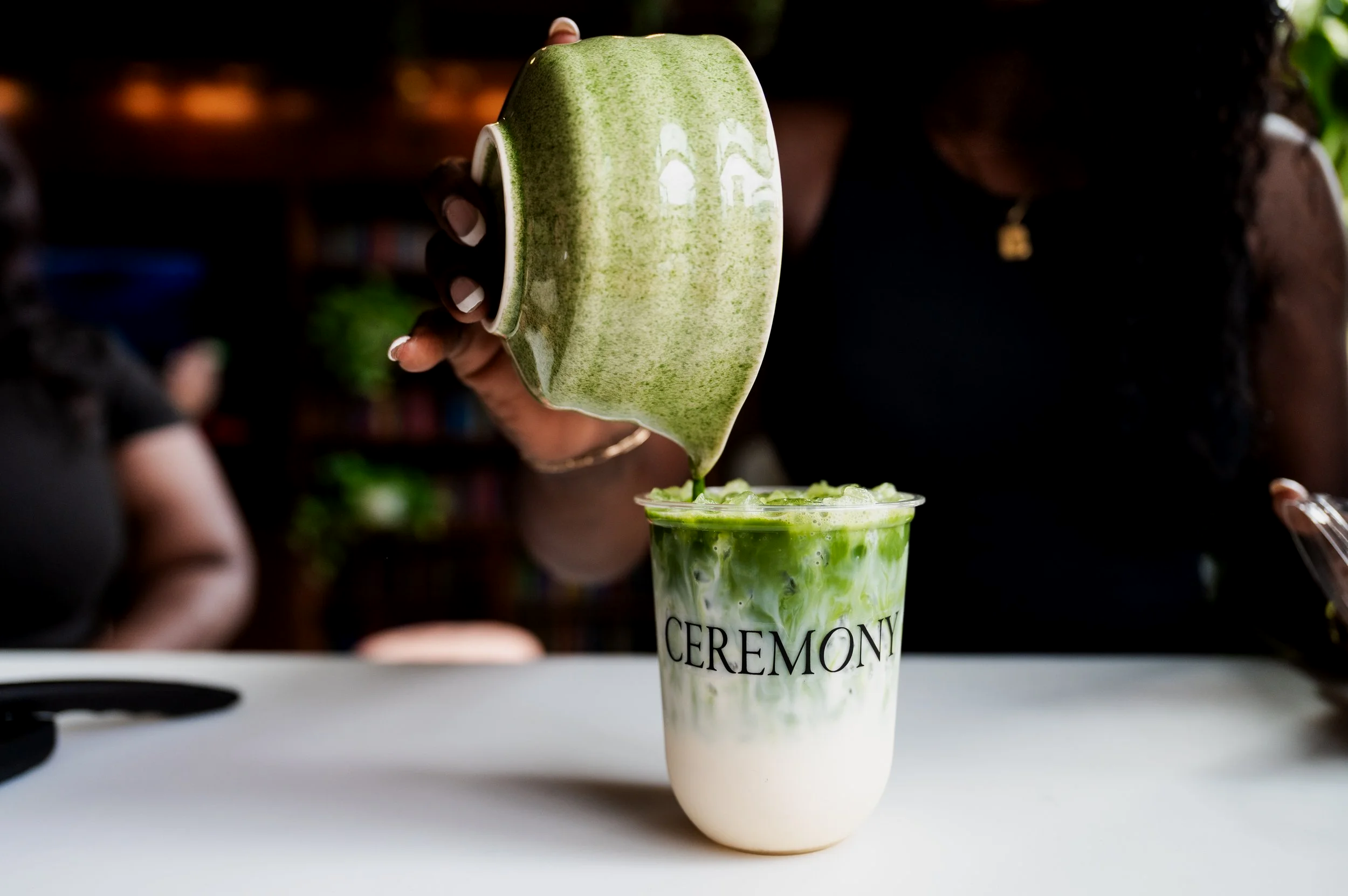 Person pouring green matcha latte from a green ceramic chawan into a clear plastic cup with the word 'CEREMONY' on it.