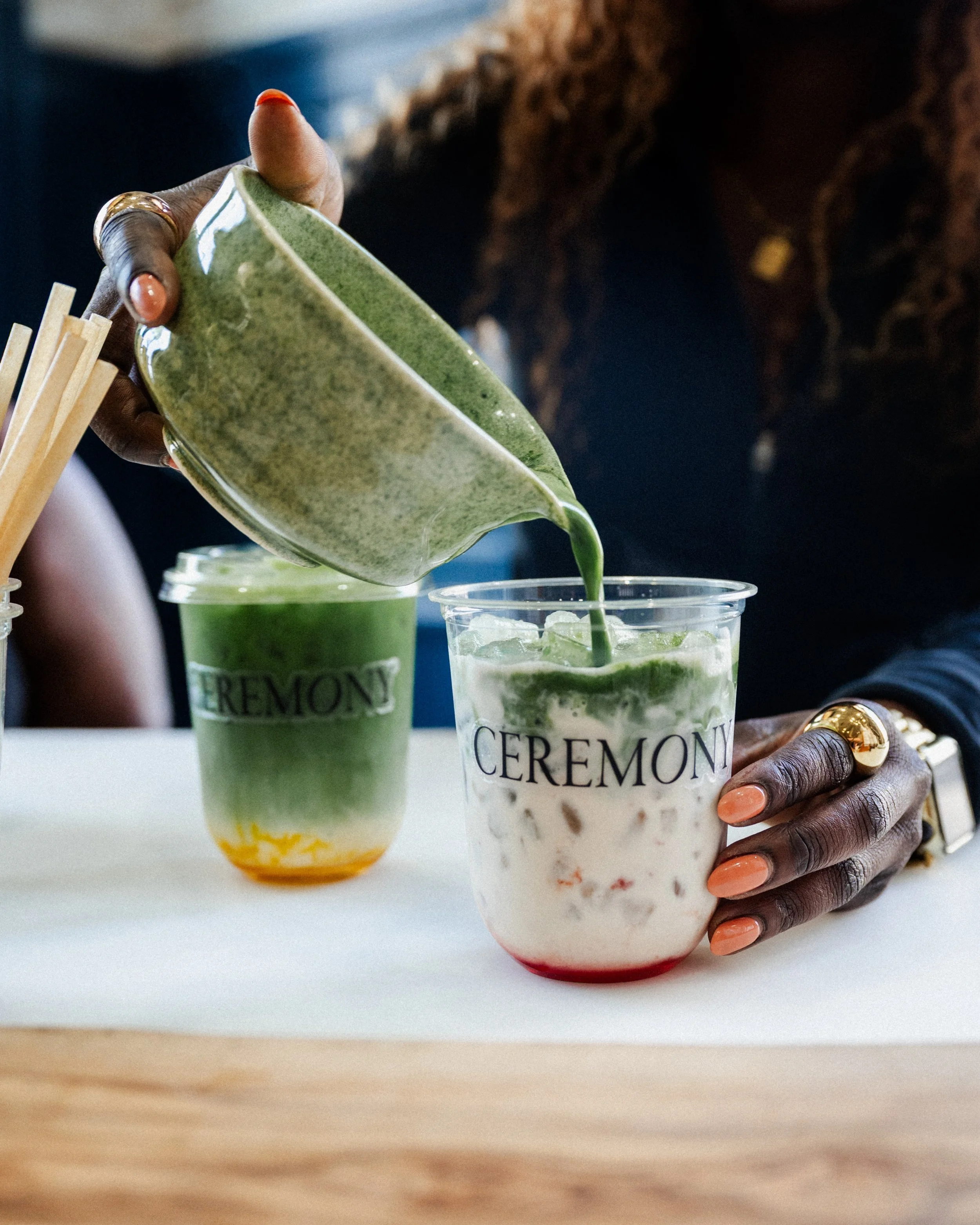 A person pours green matcha tea into a cup of iced milk with ice, sitting on a white surface, with another similar drink in the background.