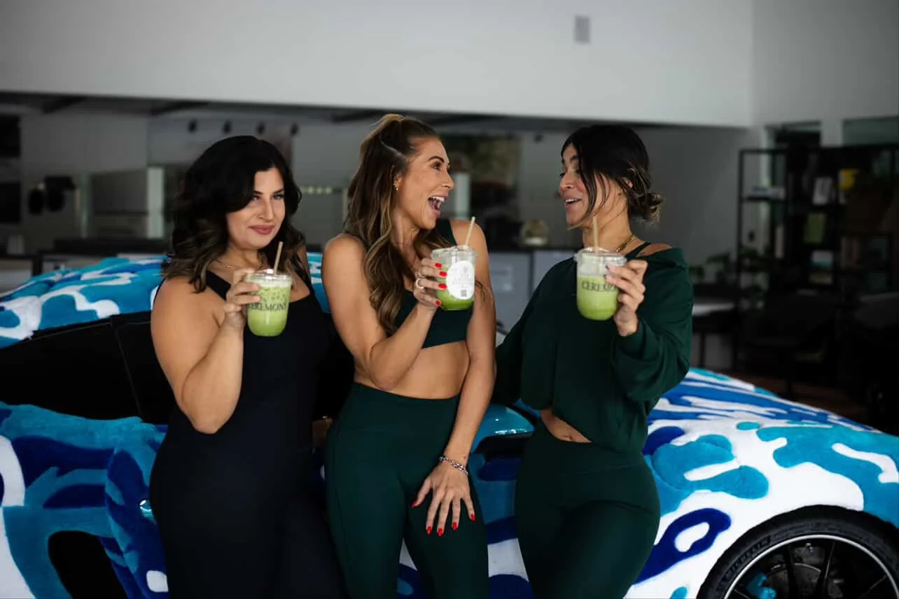 Three women standing in front of a decorated blue and white car, holding cups of matcha, and engaging in conversation.