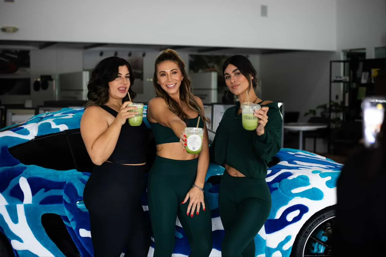 Three women standing in front of a decorated car, holding green drinks, smiling at a camera.