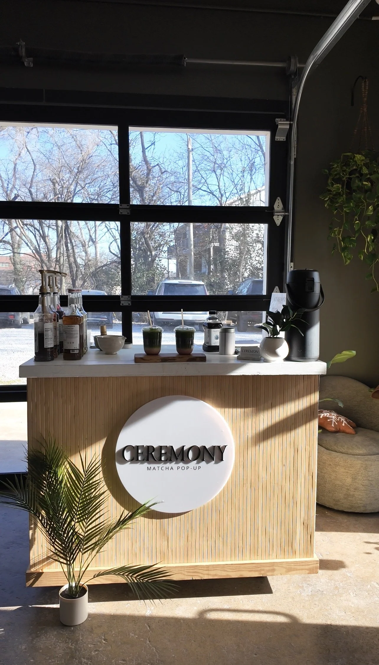 Interior of a matcha cafe with a wooden counter displaying bottles and cups, a potted plant in front, a sign reading 'CEREMONY MATCHA POP-UP', and outdoor cars visible through a large window.