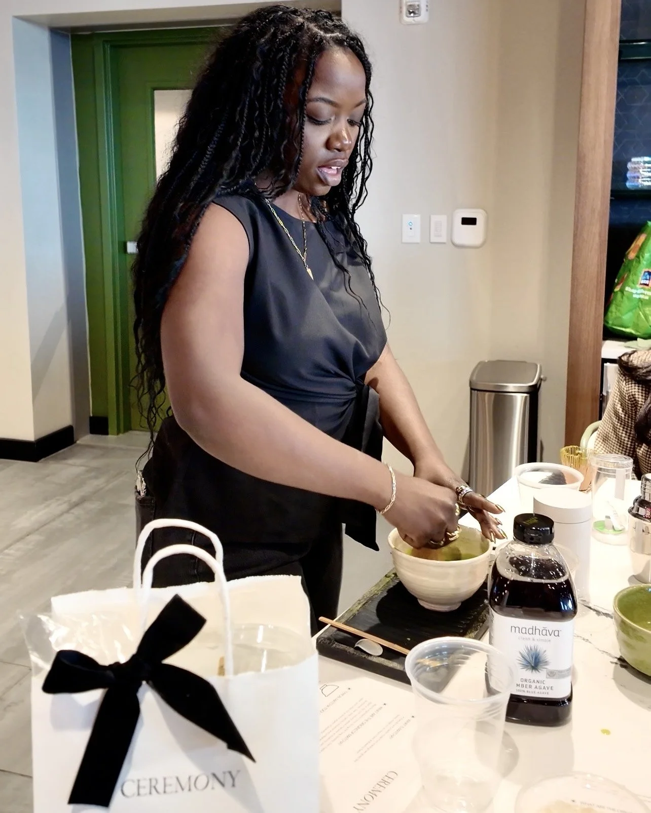 Woman preparing food or ingredients at a table during a ceremony, surrounded by various items including a white gift bag with a black bow labeled 'CEREMONY' and bottles of organic beverages.