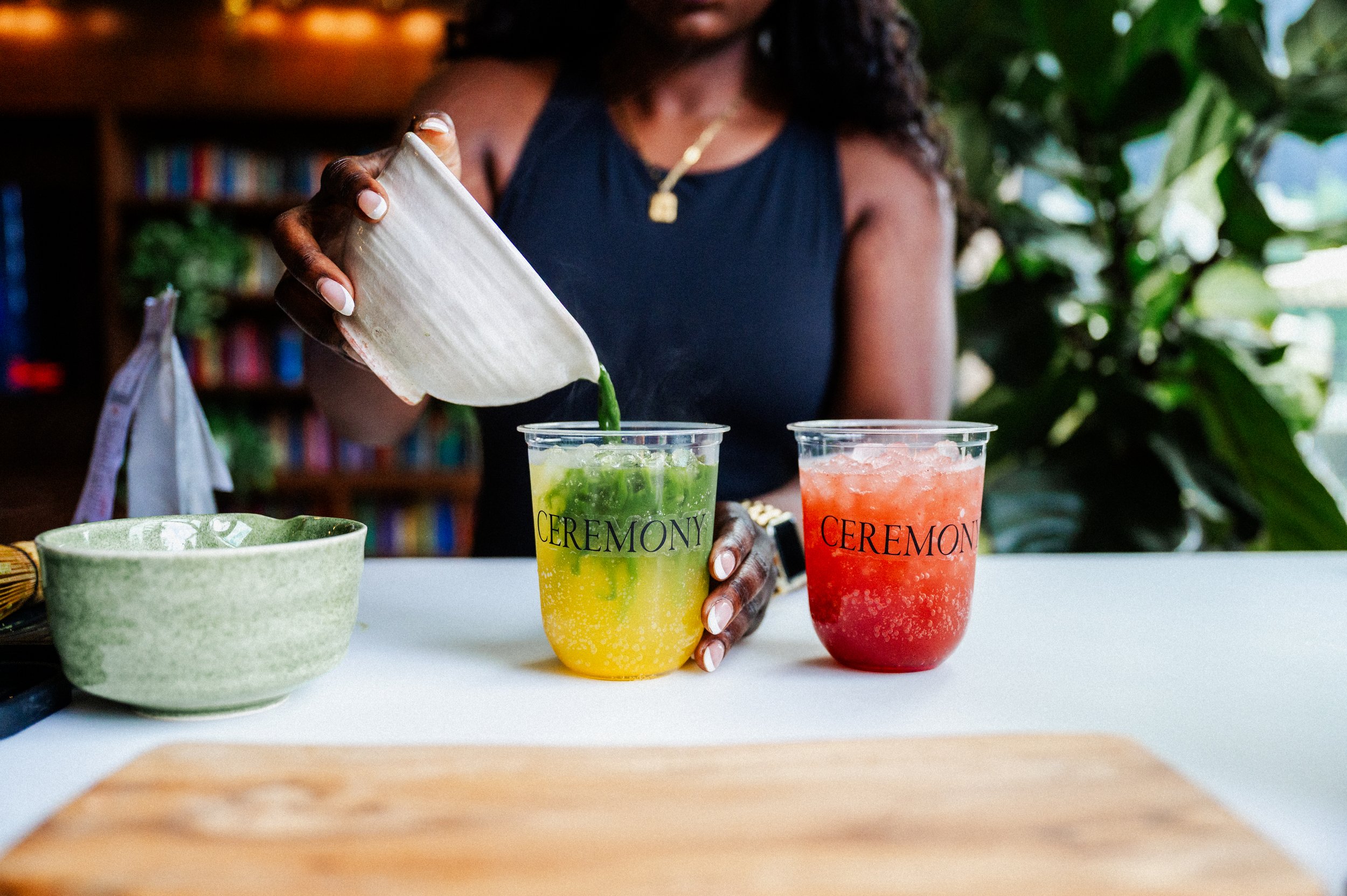 Woman pouring a green drink into a clear plastic cup labeled 'CEREMONY,' with another red drink in a similar cup also labeled 'CEREMONY,' on a table with a green bowl and a wooden cutting board, surrounded by greenery.